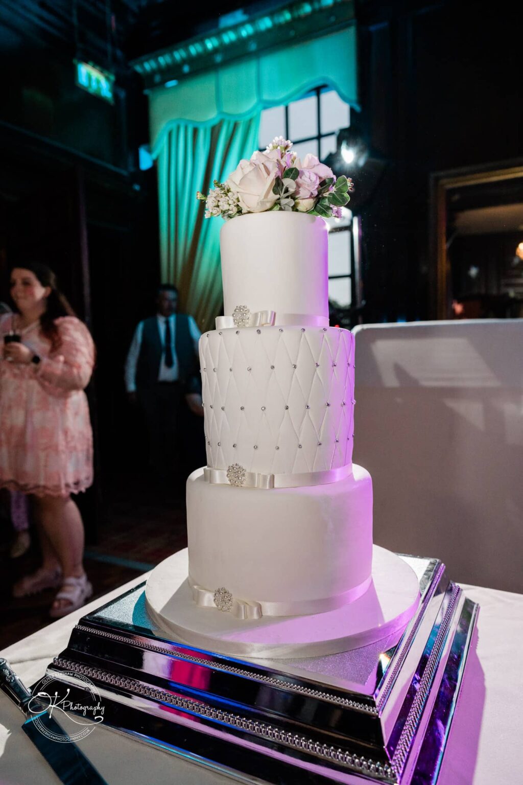 A three-tiered white wedding cake adorned with flowers and ribbon decorations, displayed on a silver cake stand in a dimly lit room.