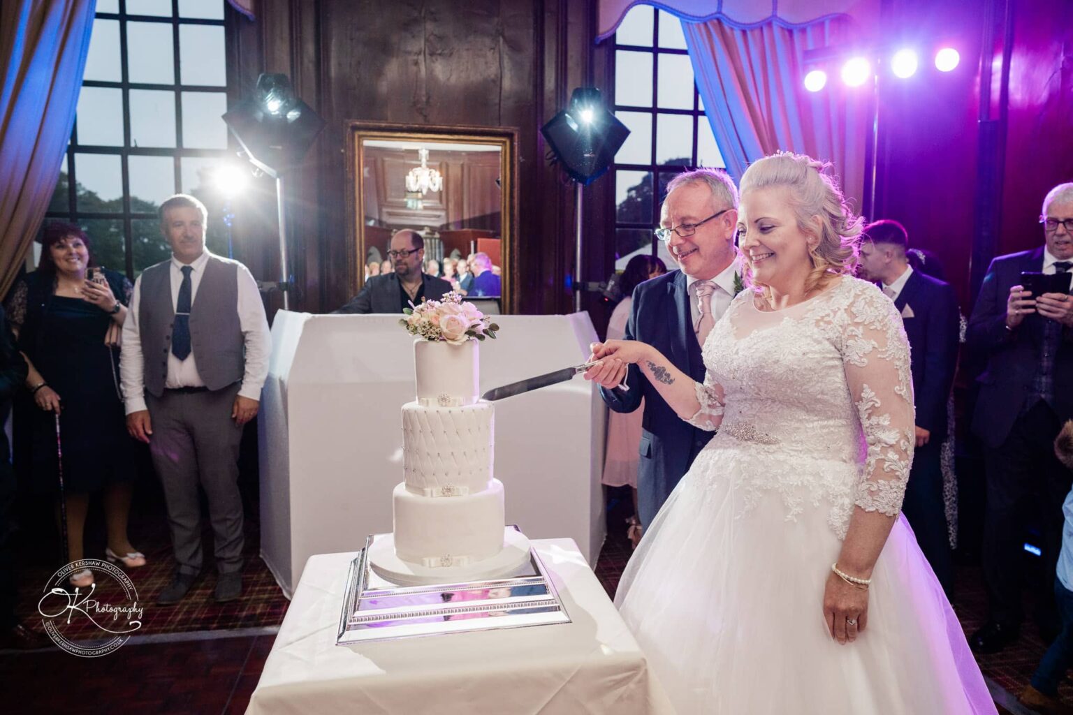 A bride and groom cutting a white three-tier wedding cake with a floral topper, surrounded by guests in an elegant venue with large windows and wooden paneling.