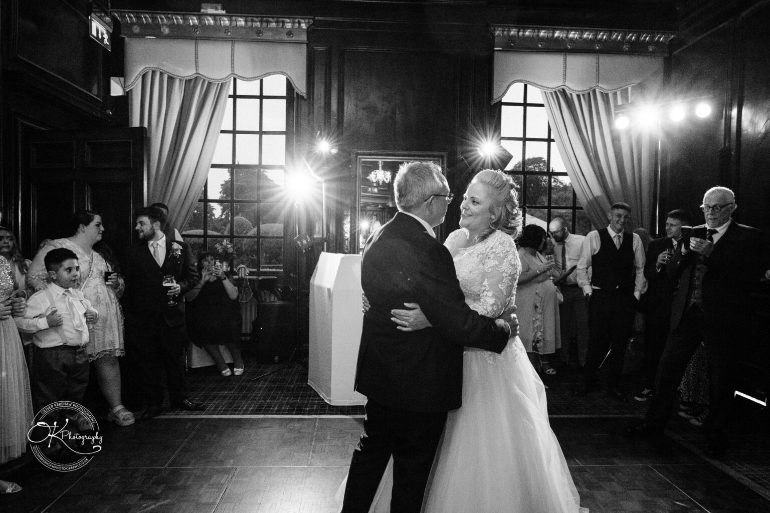 A couple dressed in wedding attire dance closely together in a dimly lit room, surrounded by smiling onlookers at Coombe Abbey Hotel.