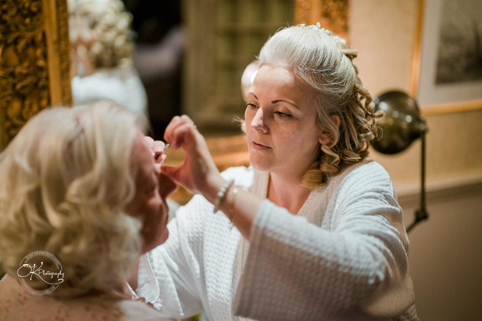 A woman applying makeup to another woman at a well-lit dressing table in a luxurious setting.