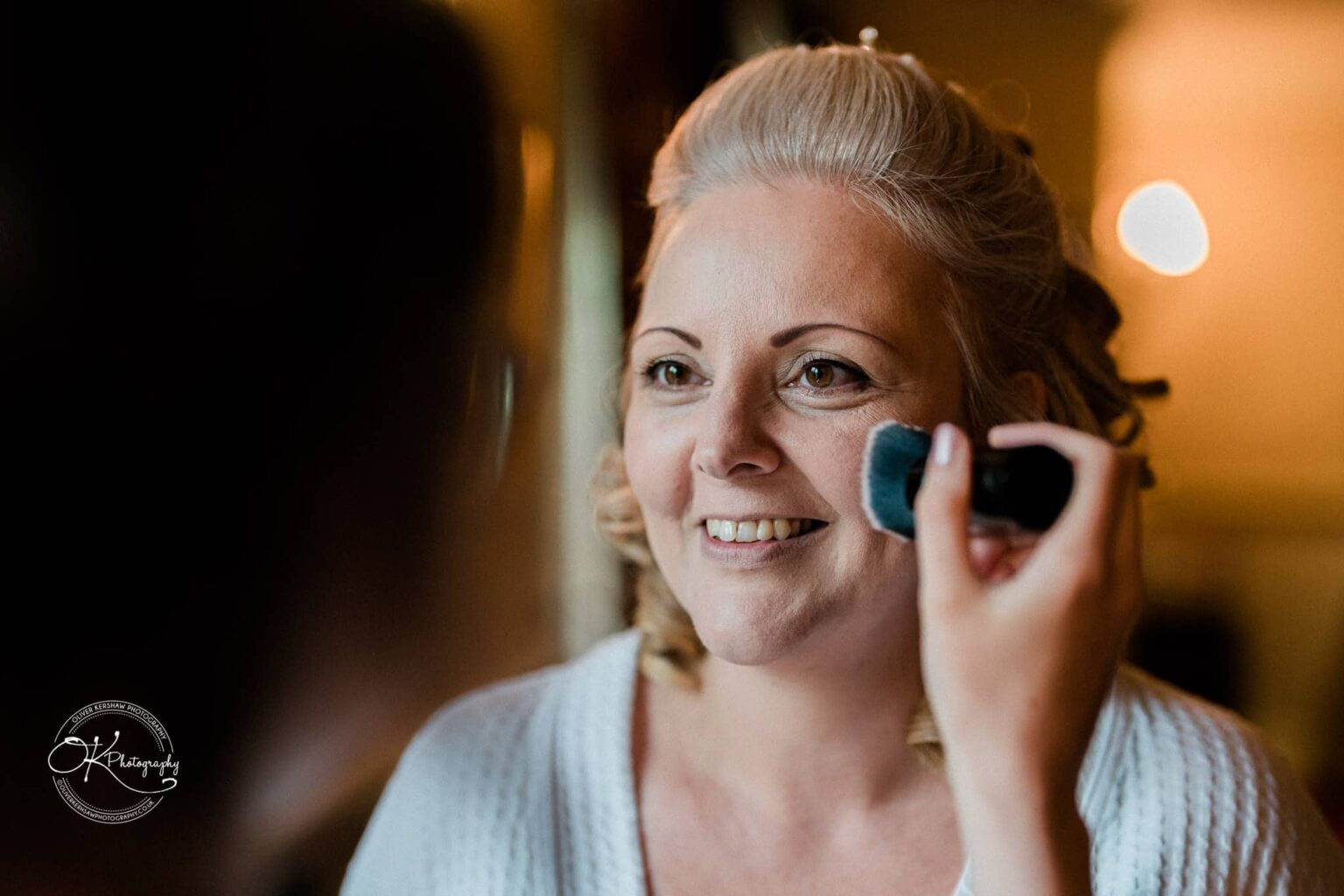A woman smiling while having makeup applied to her face.