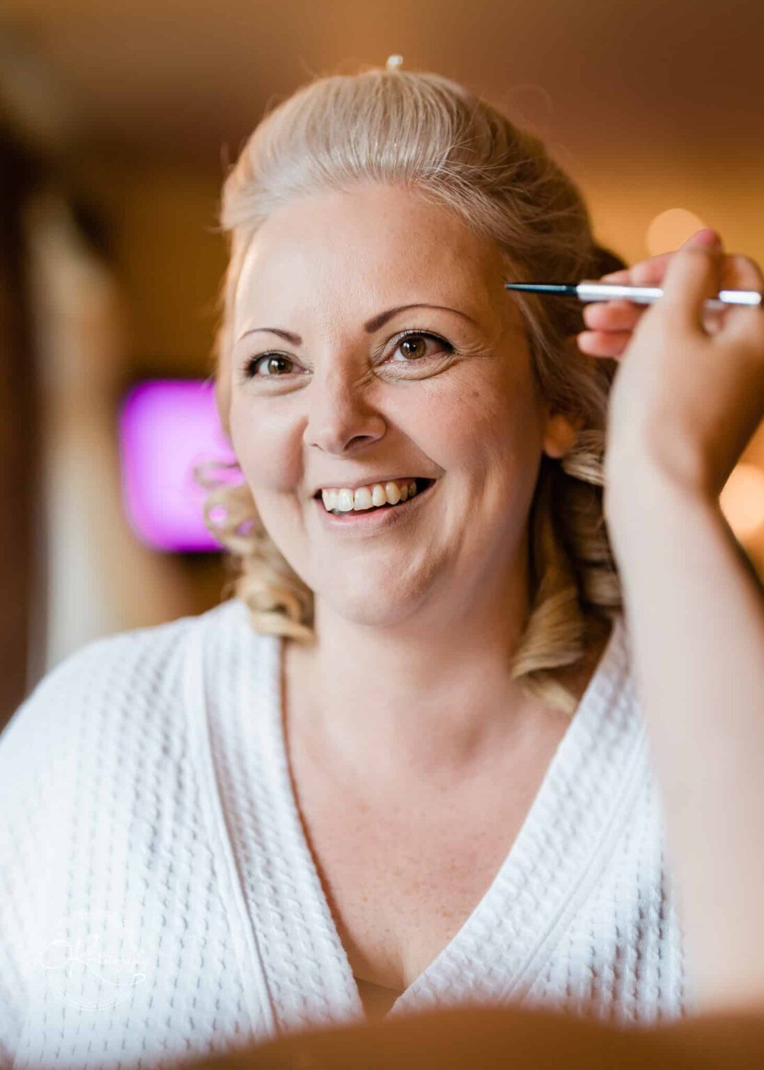 A woman smiling as she gets her makeup done, wearing a white robe.
