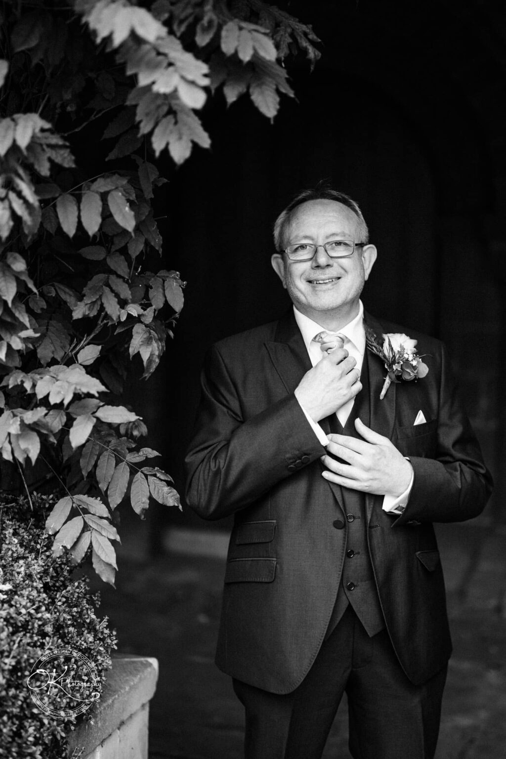 A man in a suit smiling, adjusting his tie, stands near foliage at Coombe Abbey Hotel.