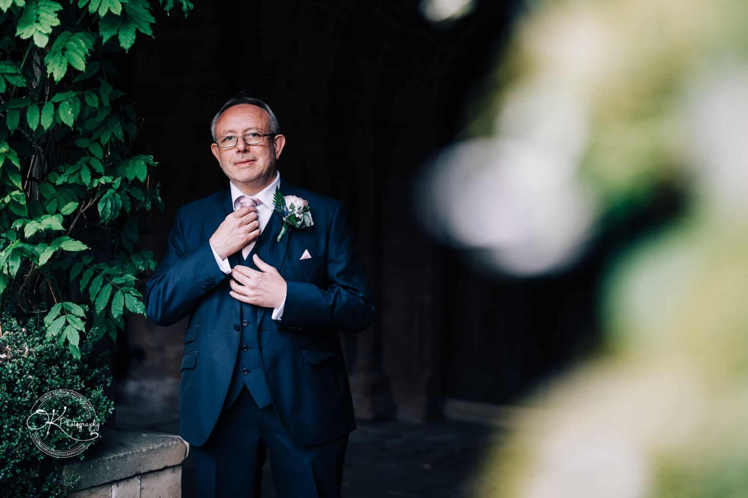 A man in a dark suit adjusts his tie, standing near greenery and smiling.
