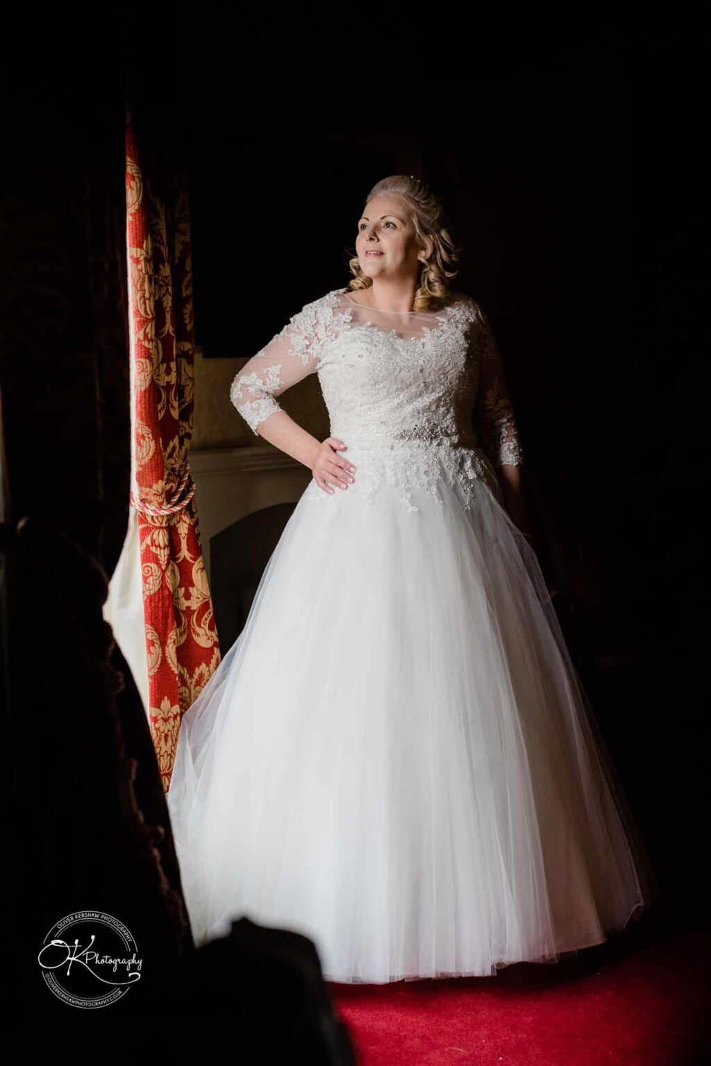 A bride in a white lace wedding dress stands indoors by a red and gold curtain with her hand on her hip, looking into the distance.