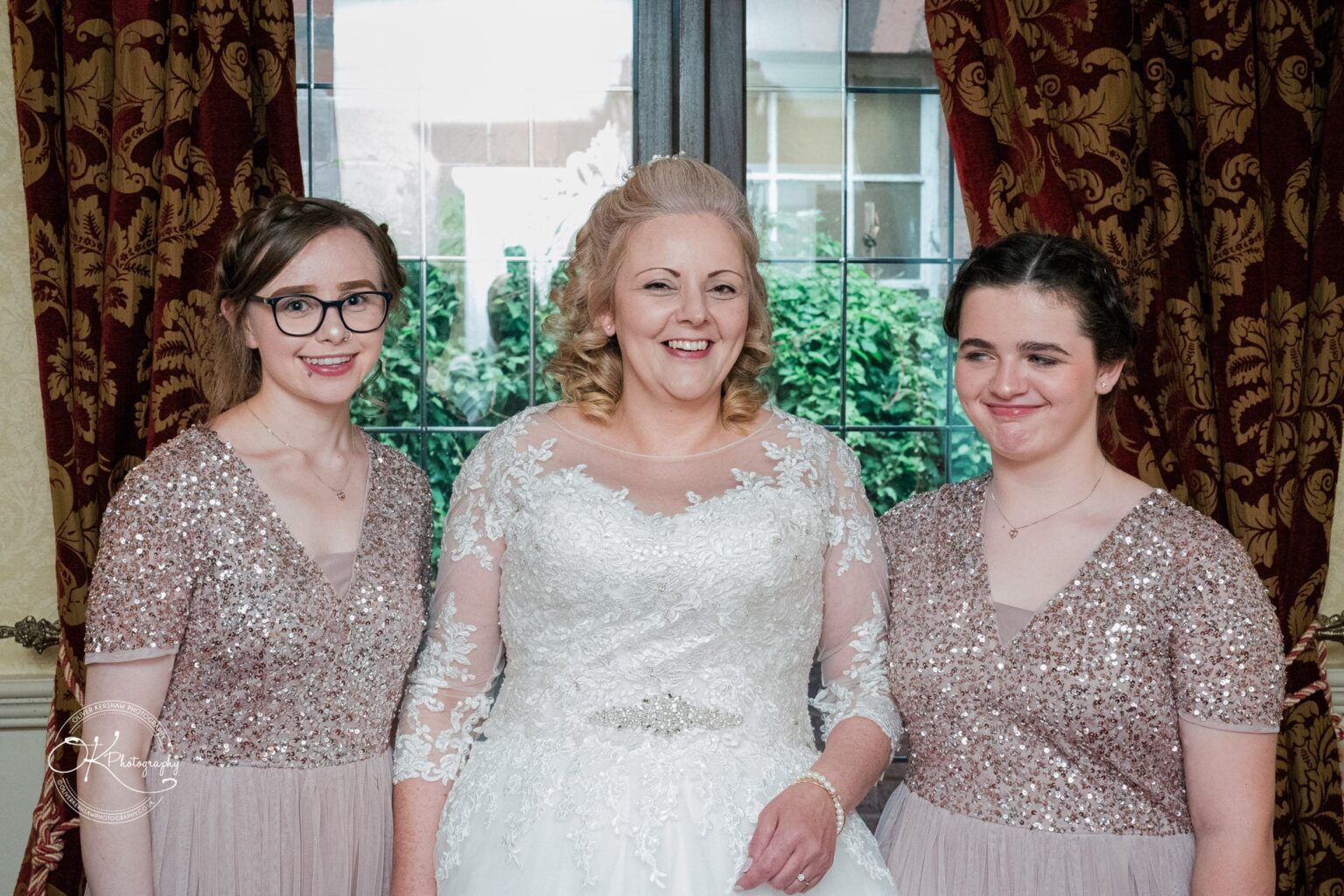 Three women standing in front of a window with patterned curtains, the middle one in a bridal gown, flanked by the other two in matching sequined dresses.