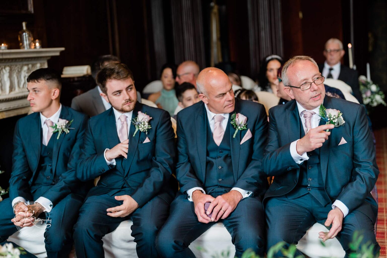Four men dressed in matching blue suits and pink ties sit together in a formal setting, with floral boutonnieres on their lapels.