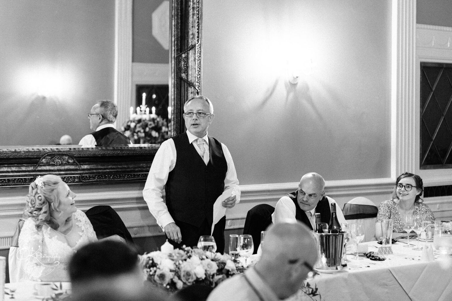 A man in a waistcoat giving a speech at a banquet table in Coombe Abbey Hotel, with wedding guests seated around him and a large mirror on the wall.