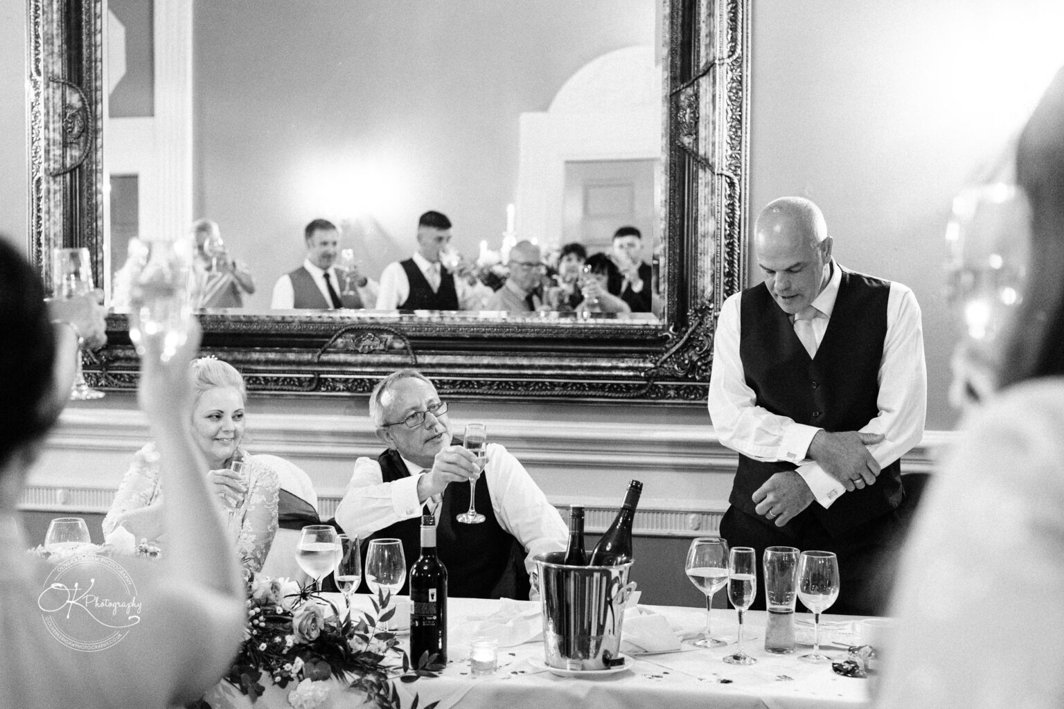 A man giving a speech at a wedding party while guests raise glasses for a toast, set in an ornate room with a large mirror.