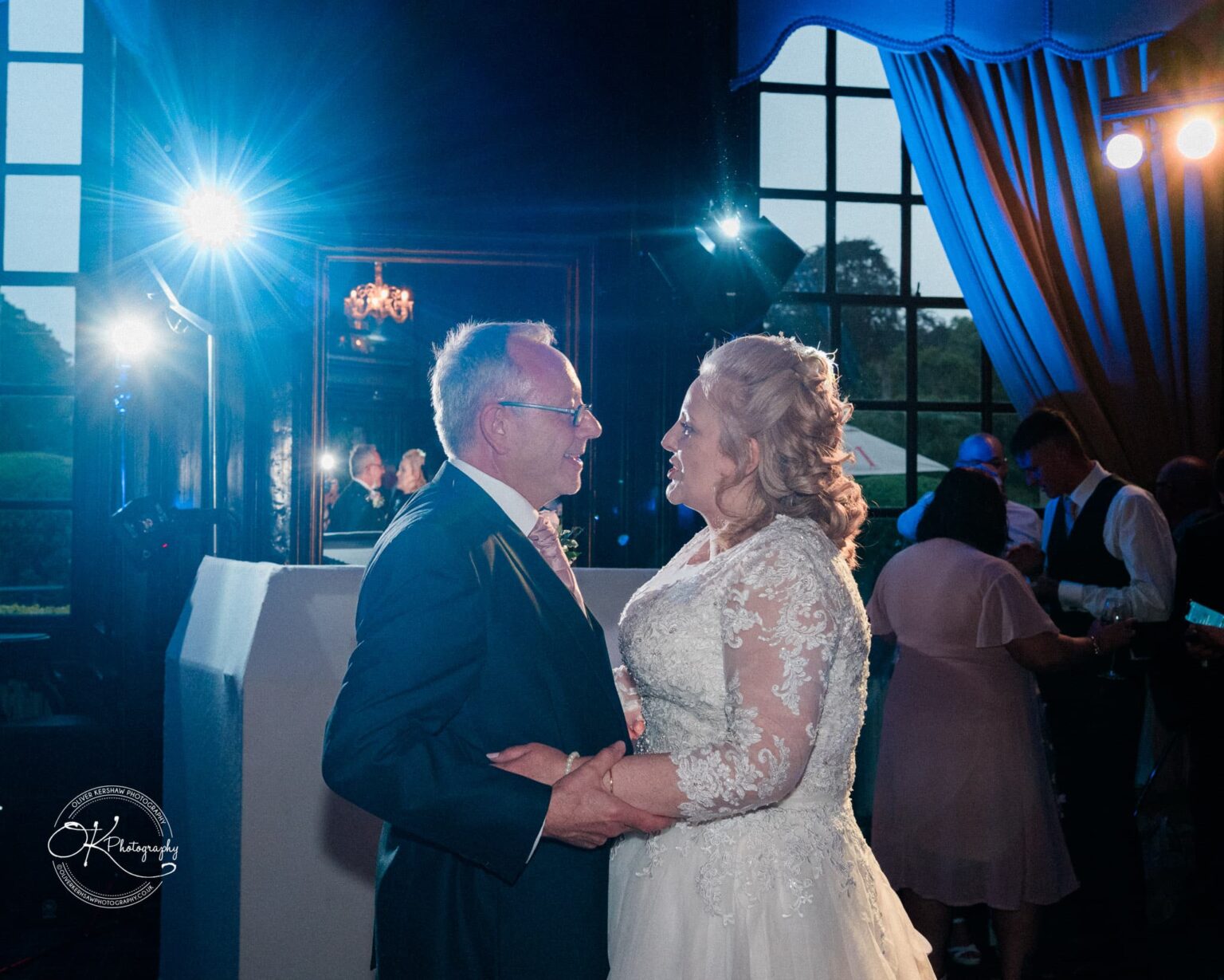 A bride and groom dancing together in a dimly lit room with guests and large windows in the background.