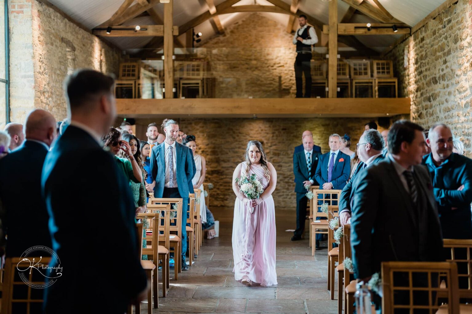 Dodford Manor Barn Wedding Photography A bridesmaid in a pink dress holding a bouquet walks down the aisle of a stone barn venue, filled with seated guests, and a photographer stands on a mezzanine above.