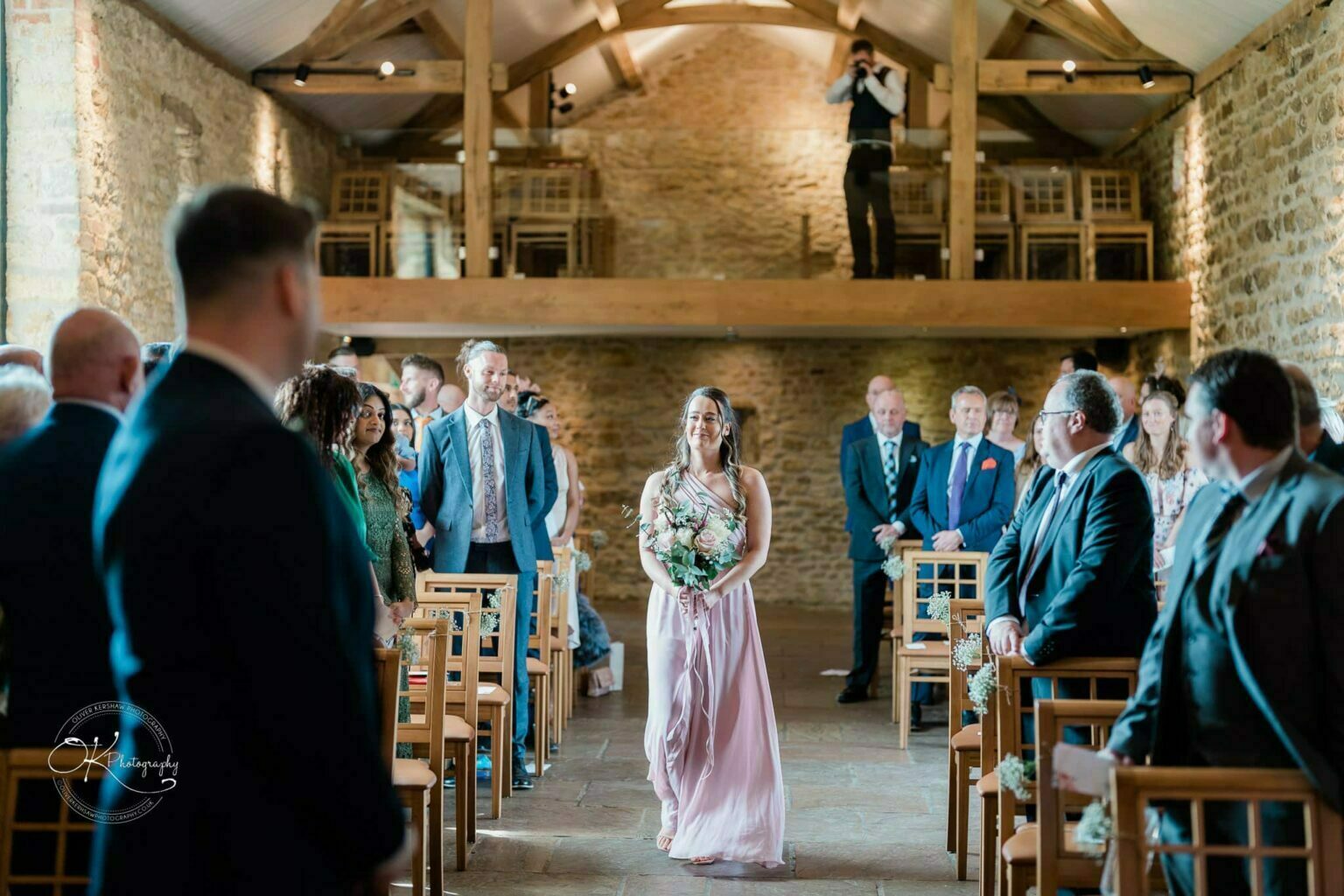 Dodford Manor Barn Wedding Photography A bridesmaid in a pink dress walks down the aisle of a decorated barn venue filled with seated guests.