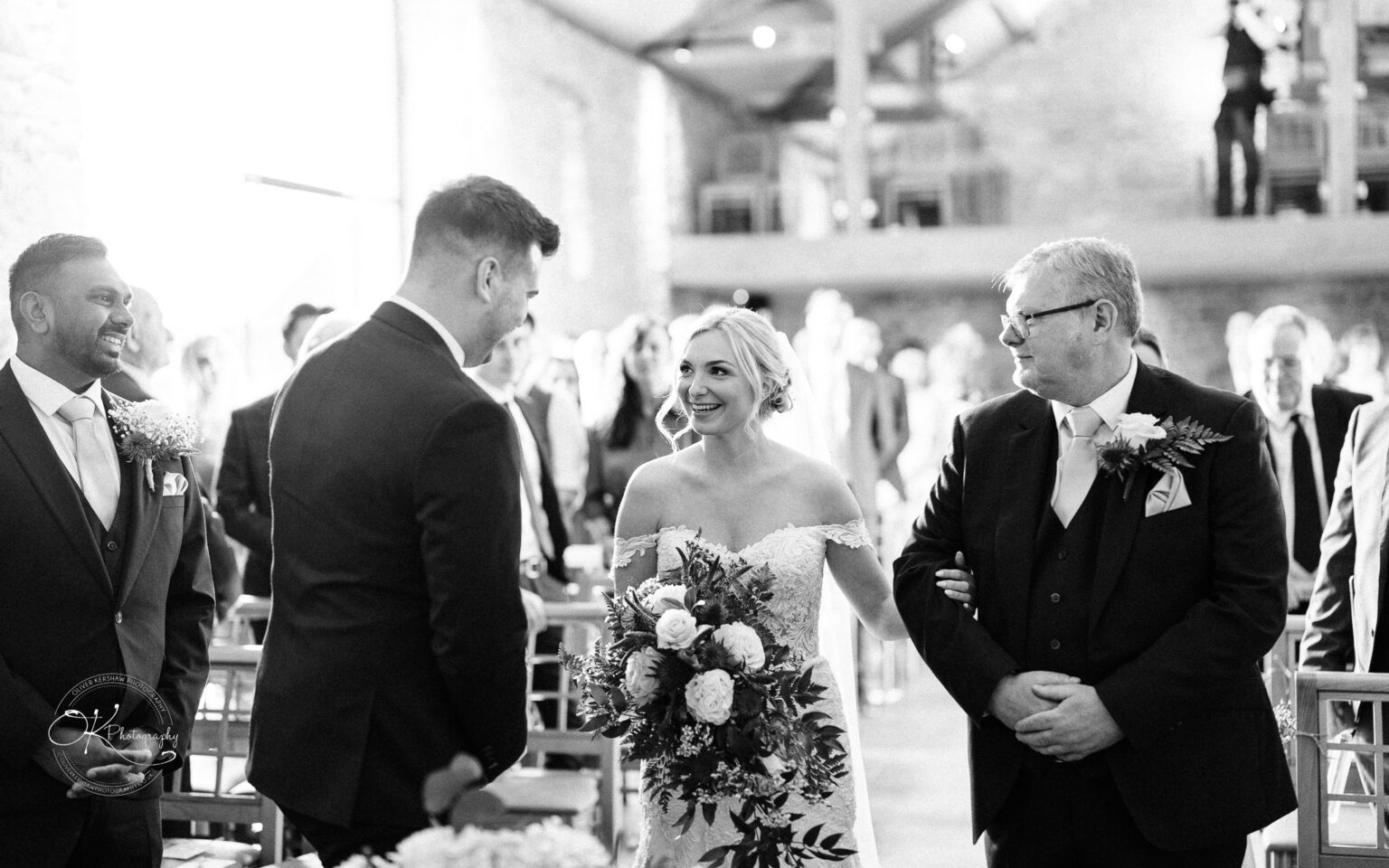 Dodford Manor Barn Wedding Photography Bride and groom smiling at each other during their wedding ceremony in a barn, with the bride holding a bouquet and accompanied by a man in a suit.