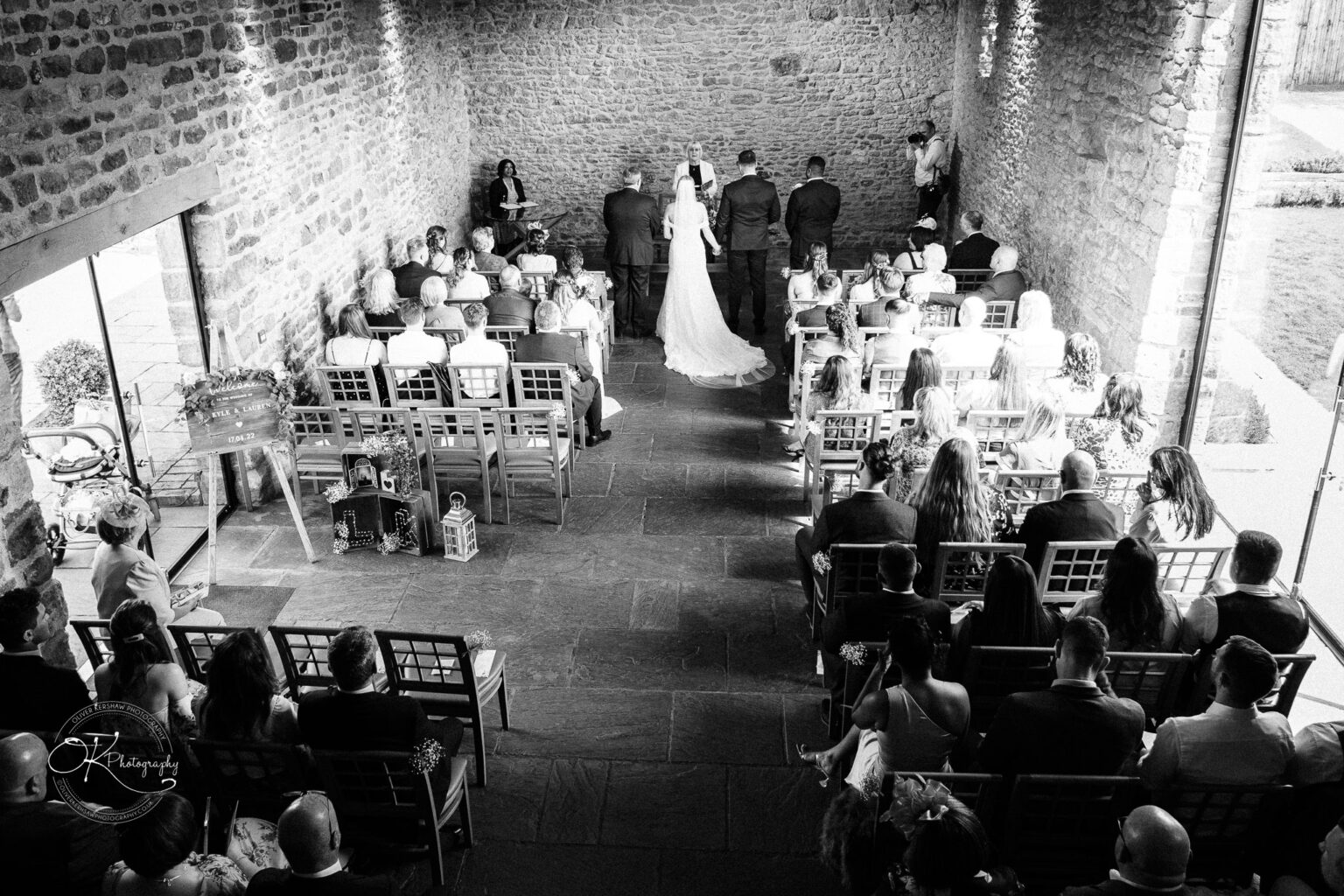 Dodford Manor Barn Wedding Photography Black and white photo of a wedding ceremony inside Dodford Manor Barn with guests seated and the couple standing at the altar.