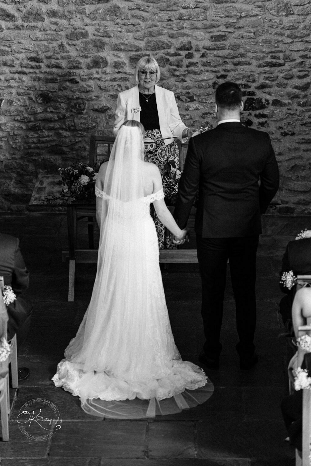 Dodford Manor Barn Wedding Photography Bride and groom holding hands and facing an officiant in a stone-walled barn.