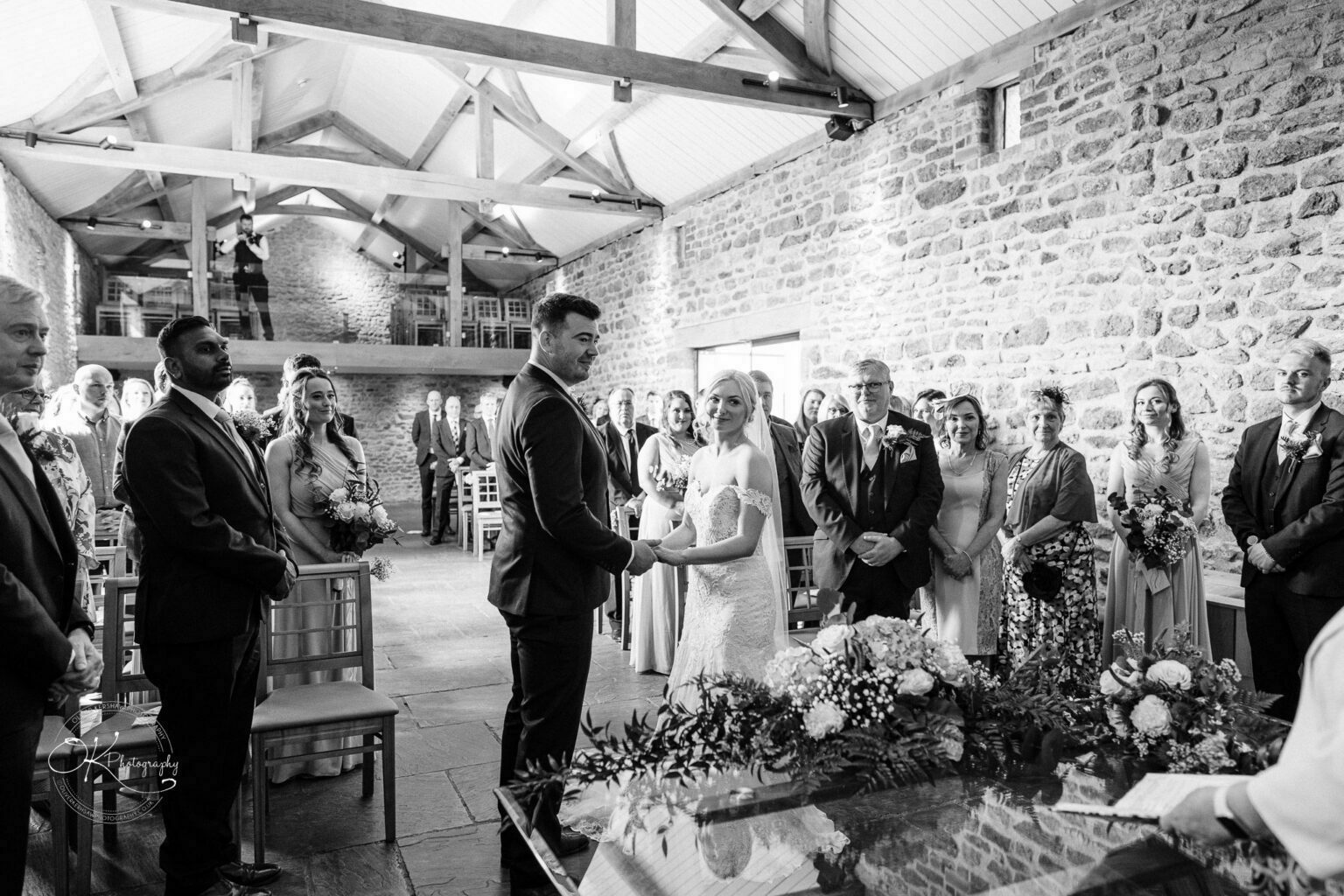 Dodford Manor Barn Wedding Photography A bride and groom standing at the altar in a barn-style wedding venue, surrounded by guests, with a photographer capturing the moment from above.