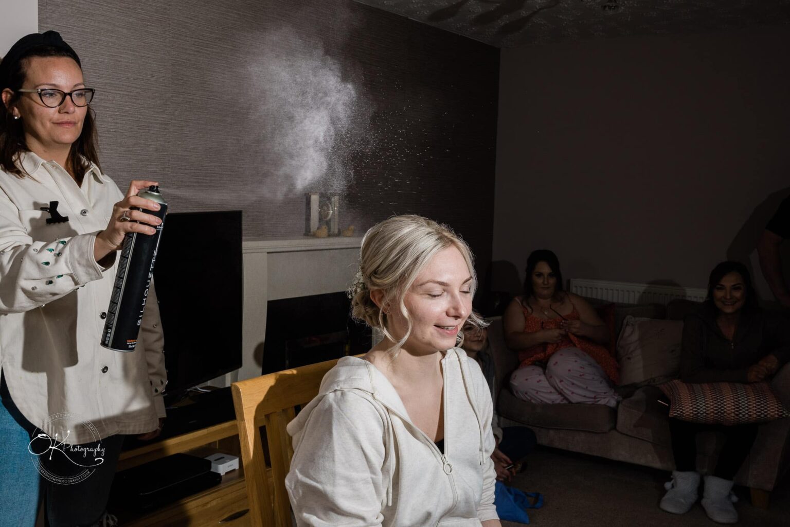 Dodford Manor Barn Wedding Photography A woman spraying hairspray on another woman's hair, with several people watching in the background.