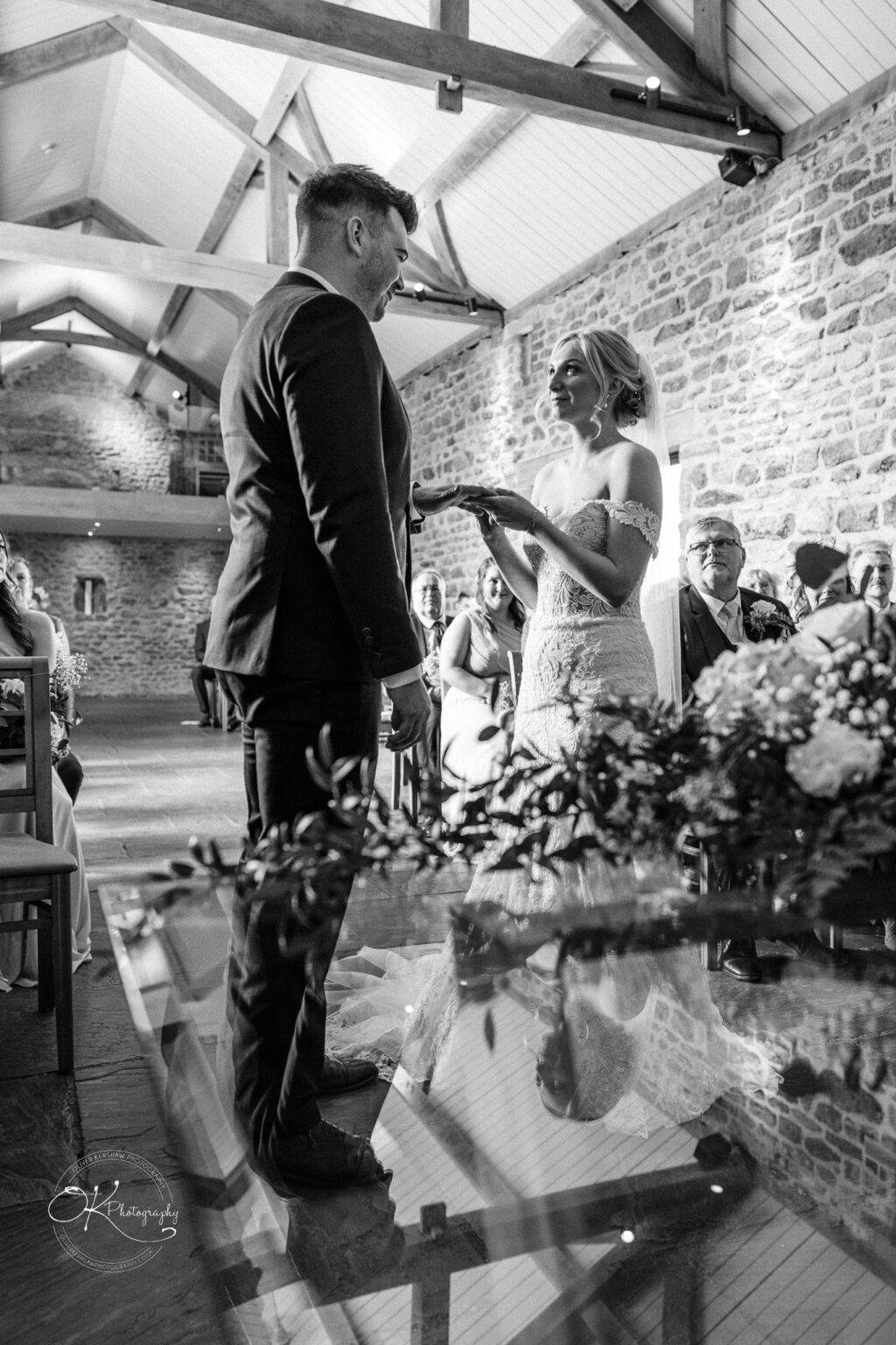 Dodford Manor Barn Wedding Photography A bride and groom exchange rings during a ceremony inside a rustic barn with exposed beams and stone walls, surrounded by seated guests.