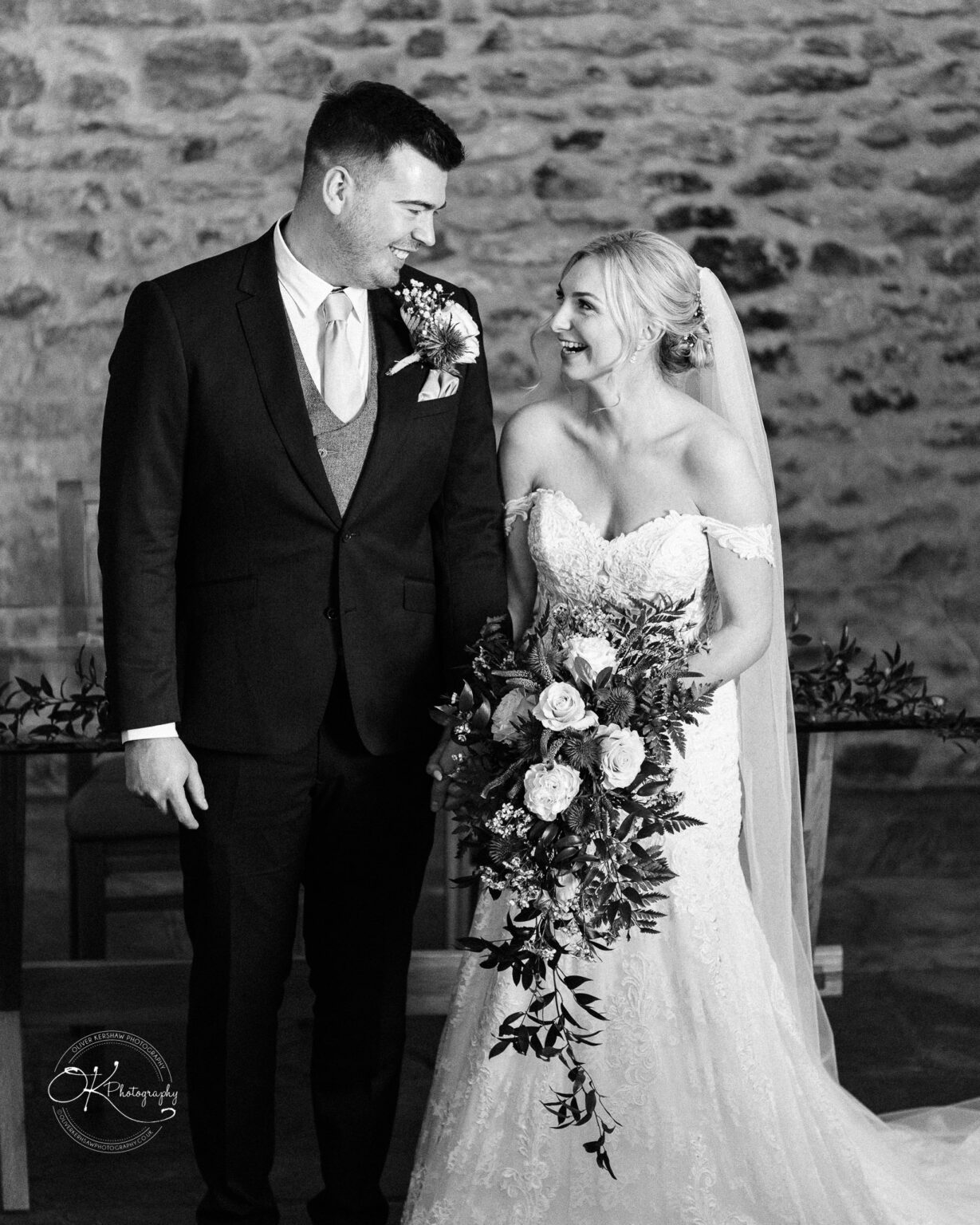 Dodford Manor Barn Wedding Photography A bride and groom smiling at each other, standing indoors in a rustic stone-walled barn. The bride is holding a bouquet of flowers.