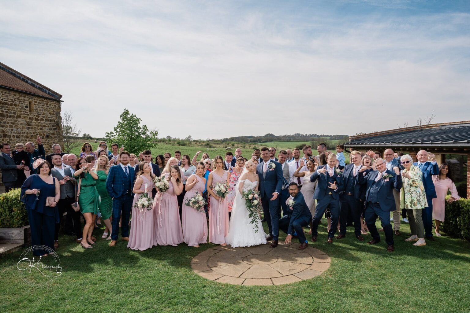 Dodford Manor Barn Wedding Photography A large wedding party posing together outdoors at Dodford Manor Barn, with the bride and groom in the center surrounded by bridesmaids in pink dresses and groomsmen in blue suits, against a rural landscape.