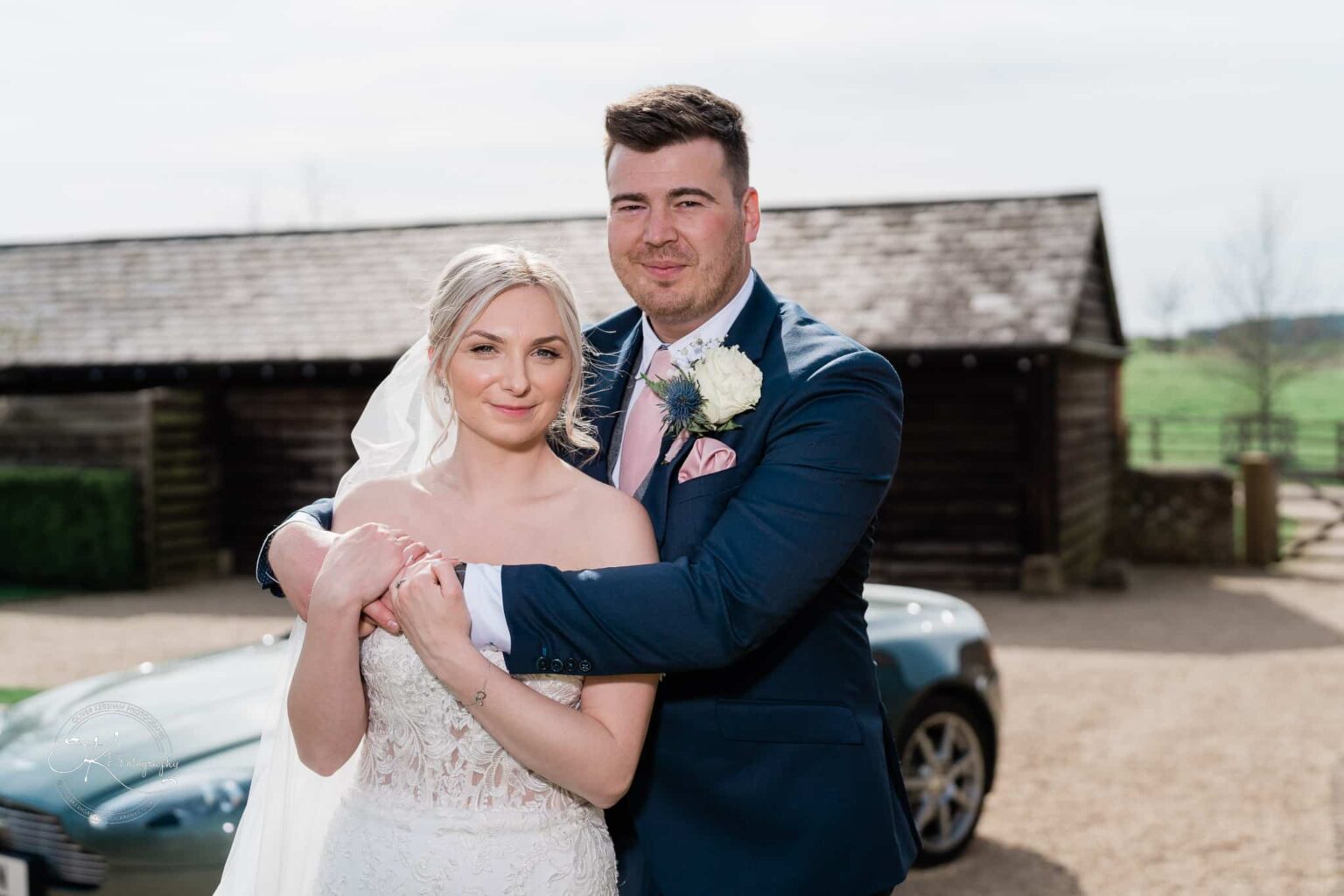Dodford Manor Barn Wedding Photography A bride and groom embrace in front of a rustic barn at Dodford Manor.