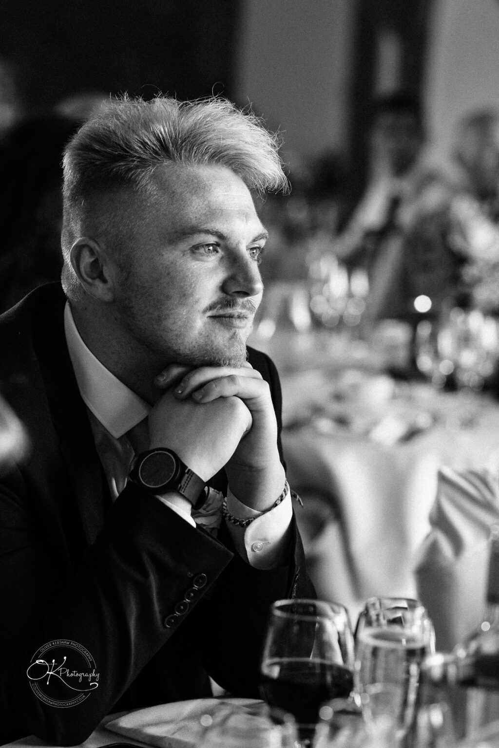 Dodford Manor Barn Wedding Photography A man in formal attire gazes thoughtfully to the side at a dining event, resting his chin on his hands, with blurred table settings in the background.