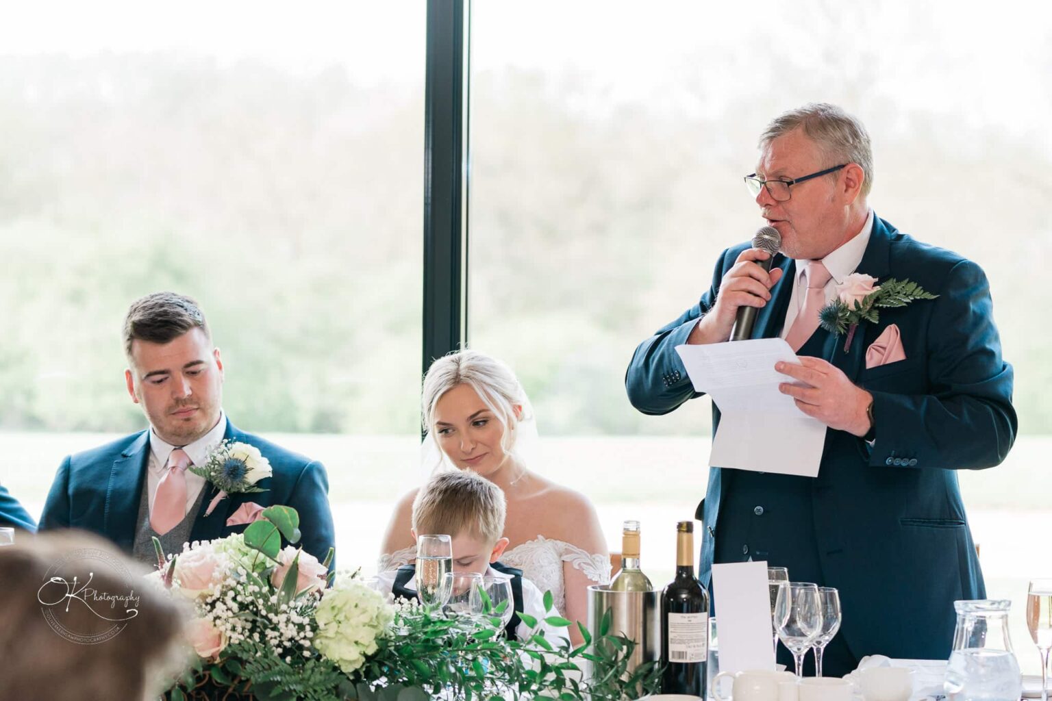 Dodford Manor Barn Wedding Photography A wedding reception at Dodford Manor Barn with a seated bride and groom listening to a man giving a speech using a microphone.