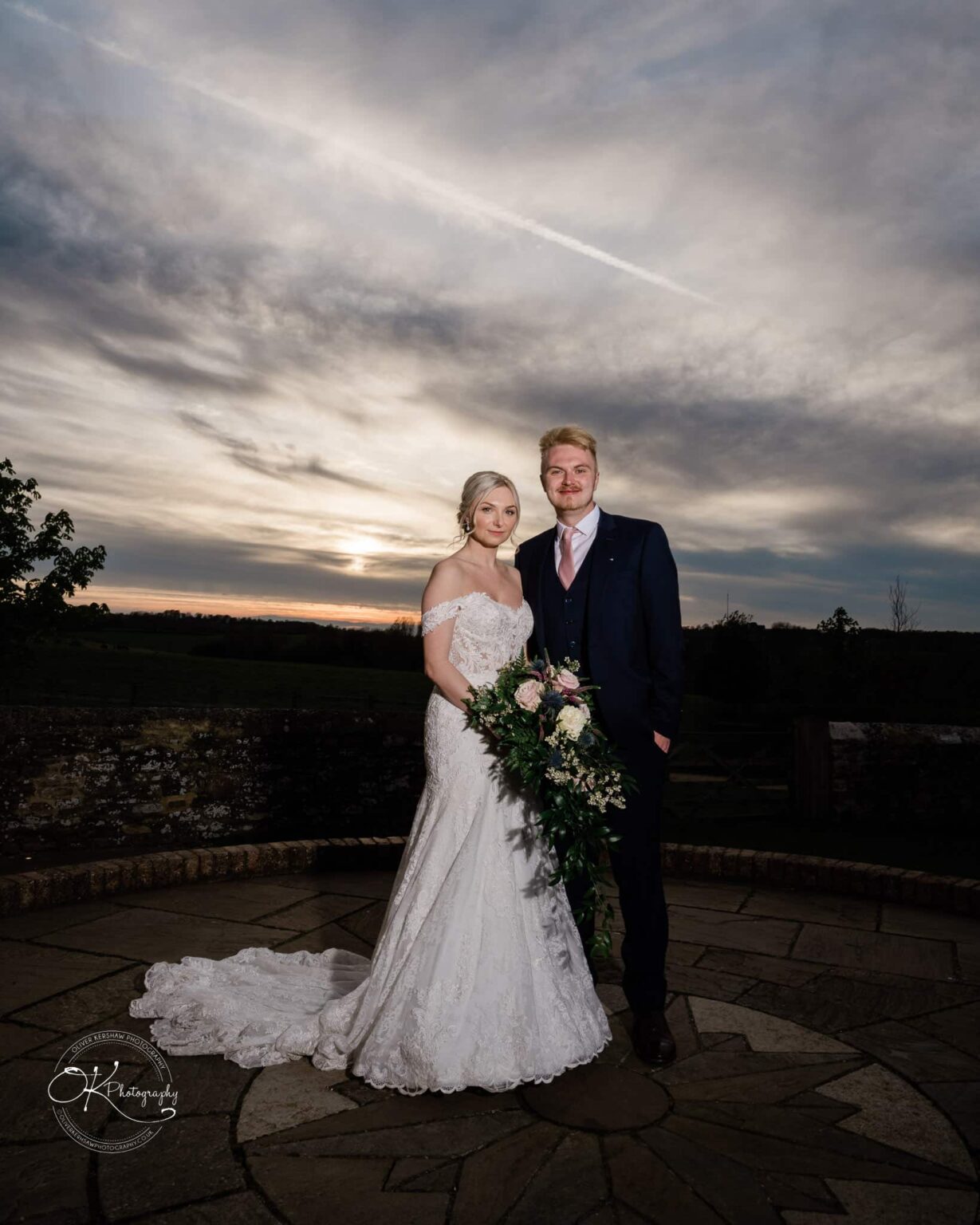 Dodford Manor Barn Wedding Photography A bride and groom standing outside on a stone patio at sunset. The bride is wearing a white lace gown and holding a bouquet, while the groom is in a dark suit with a pink tie.