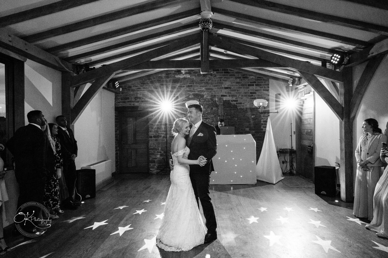 Dodford Manor Barn Wedding Photography Bride and groom share a first dance in a rustic barn setting with wooden beams and brick walls, surrounded by guests.