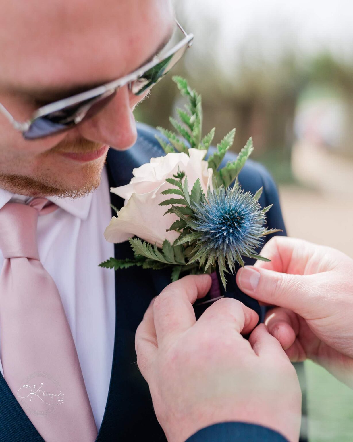 Dodford Manor Barn Wedding Photography Close-up of a man wearing sunglasses and having a boutonniere with a rose and blue thistle pinned to his suit.