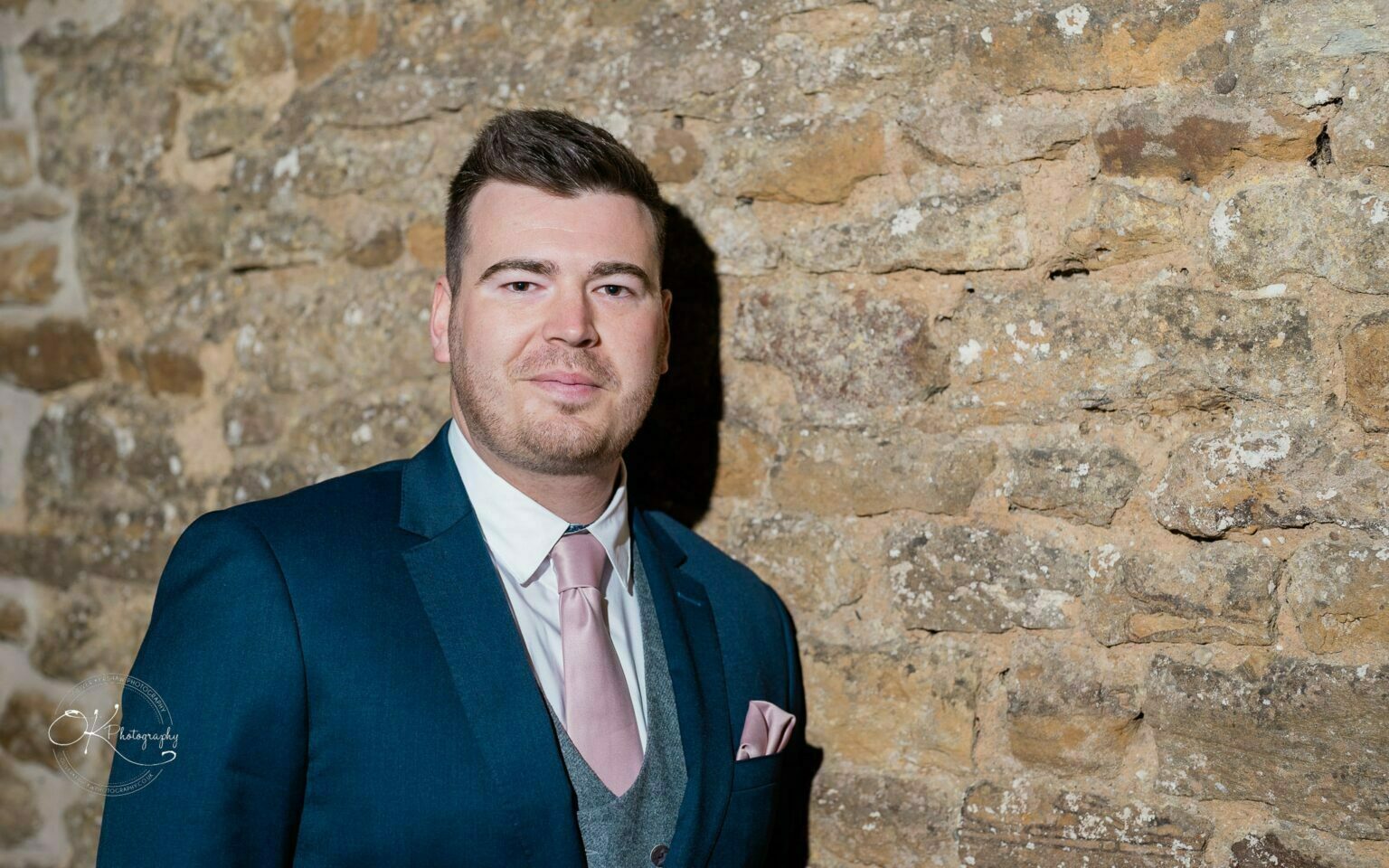 Dodford Manor Barn Wedding Photography Man in a suit and pink tie stands in front of a stone wall.