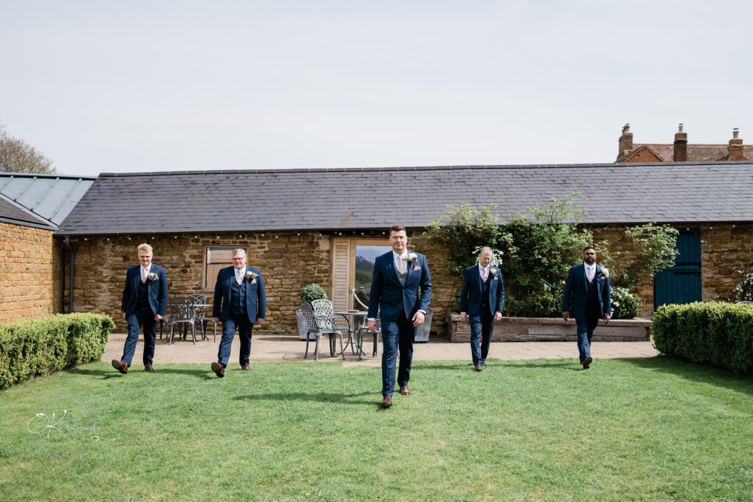 Dodford Manor Barn Wedding Photography Five men in formal attire walking across a grassy courtyard in front of a stone building at Dodford Manor Barn.