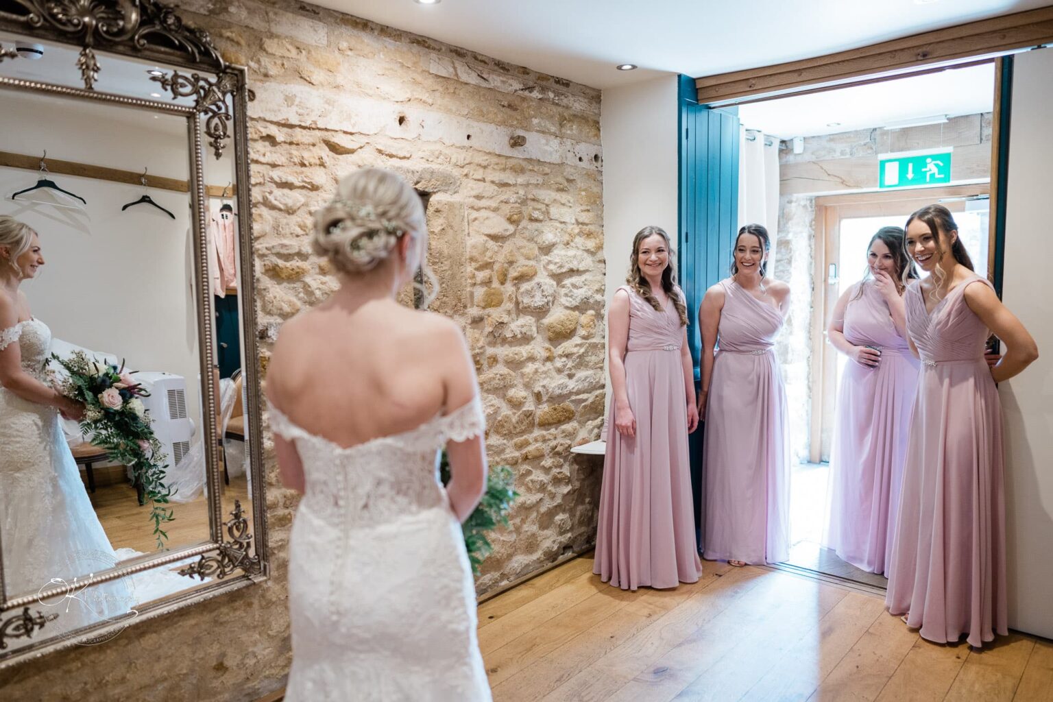 Dodford Manor Barn Wedding Photography A bride in a white gown stands in front of a mirror, with four bridesmaids in pink dresses smiling and chatting in a room with stone walls at Dodford Manor Barn