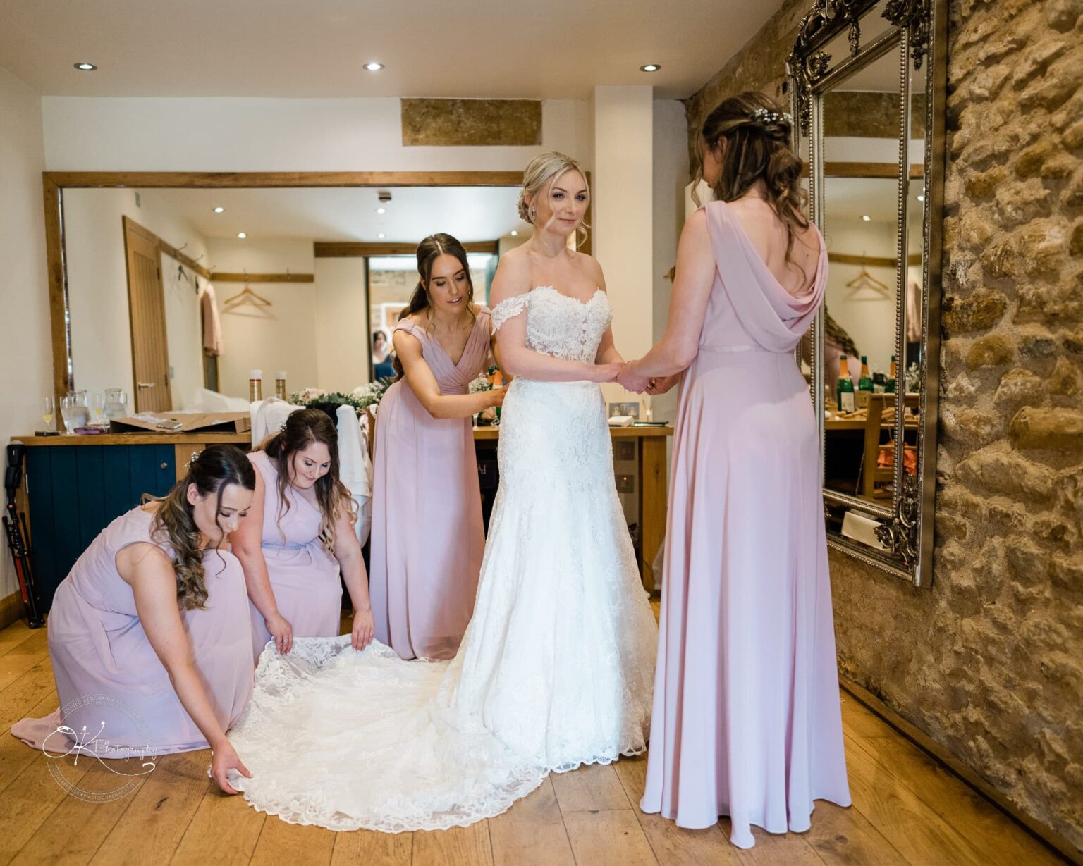 Dodford Manor Barn Wedding Photography Bride in lace wedding dress with bridesmaids in light pink dresses helping to arrange her gown train in rustic room with stone wall and wooden floor.