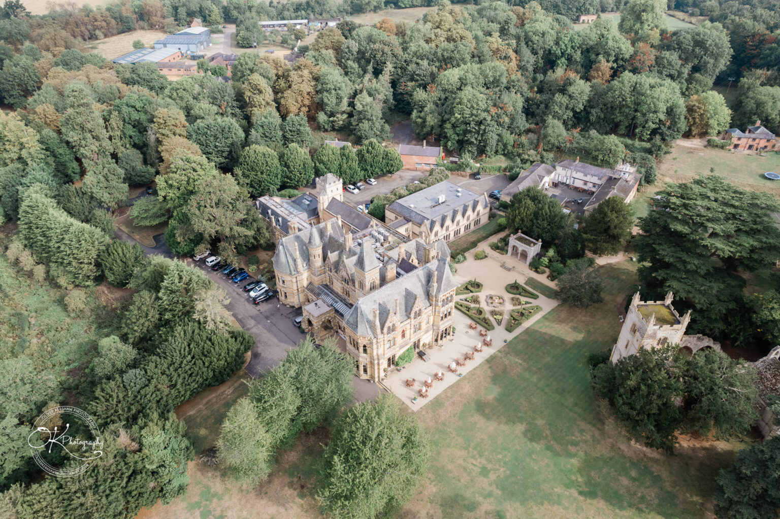 Ettington Park Hotel Wedding Photography Aerial view of a large, historic building surrounded by trees, gardens, and smaller buildings, with a spacious landscaped area and parked cars nearby.