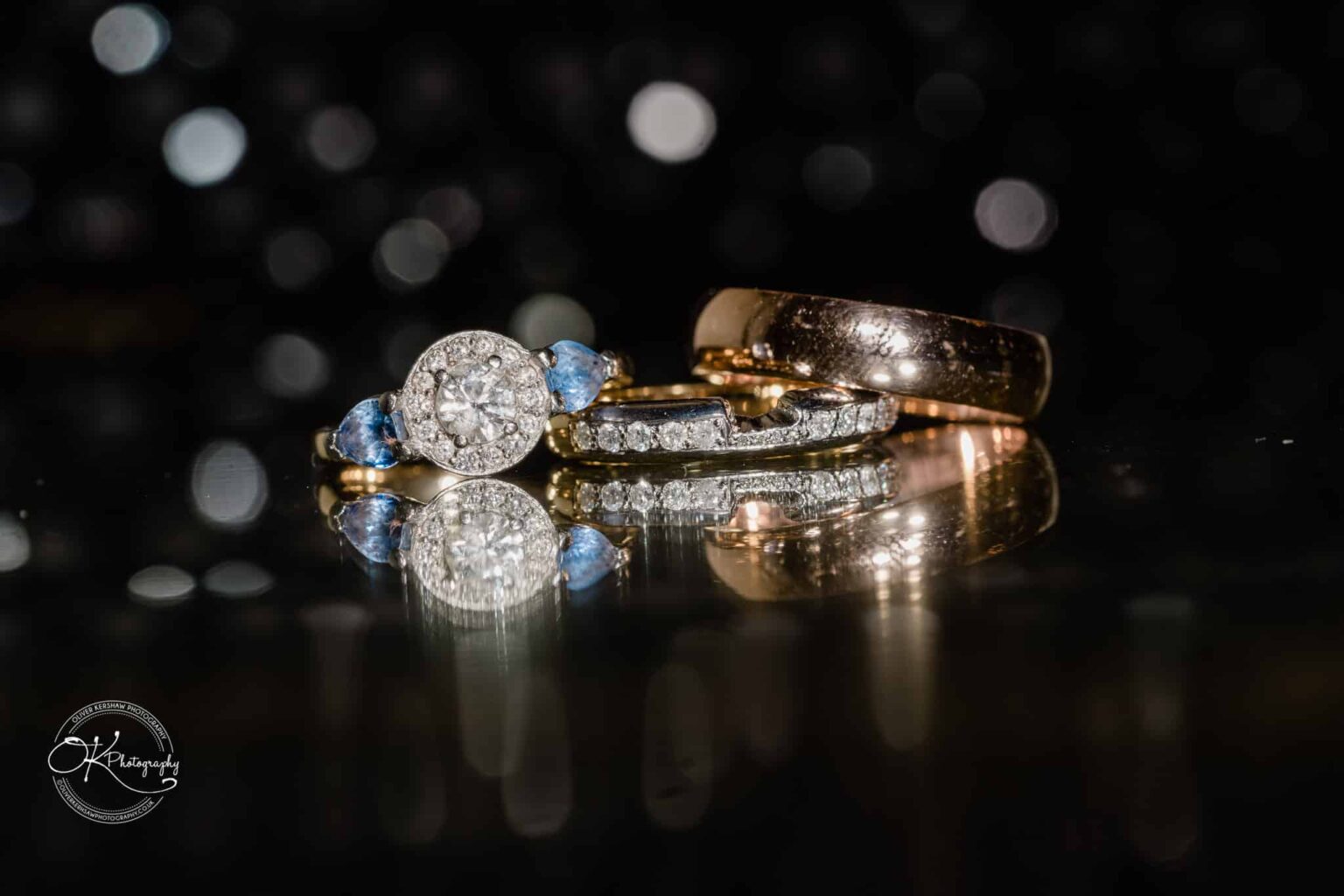 Ettington Park Hotel Wedding Photography Close-up of wedding rings and a diamond engagement ring with blue gems arranged on a reflective surface.