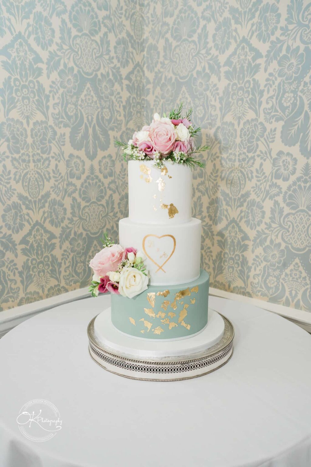 Ettington Park Hotel Wedding Photography A three-tier wedding cake with floral decorations on top of each tier, gold leaf accents, and an initialed heart design on the middle tier. The bottom tier is light blue, the top two tiers are white. The cake sits on a round table with a white tablecloth against a patterned wallpaper background.