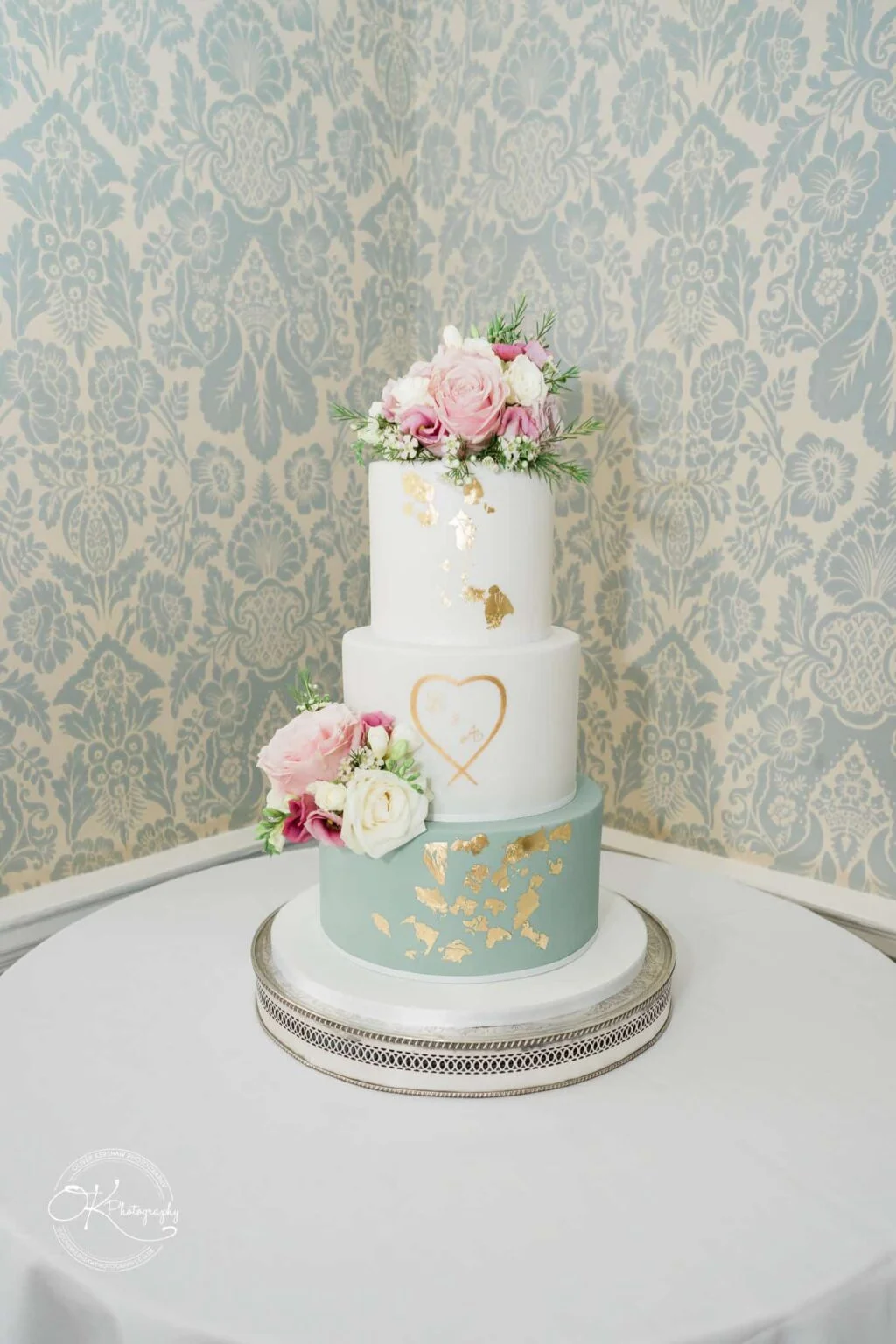 Ettington Park Hotel Wedding Photography A three-tier wedding cake with floral decorations on top of each tier, gold leaf accents, and an initialed heart design on the middle tier. The bottom tier is light blue, the top two tiers are white. The cake sits on a round table with a white tablecloth against a patterned wallpaper background.