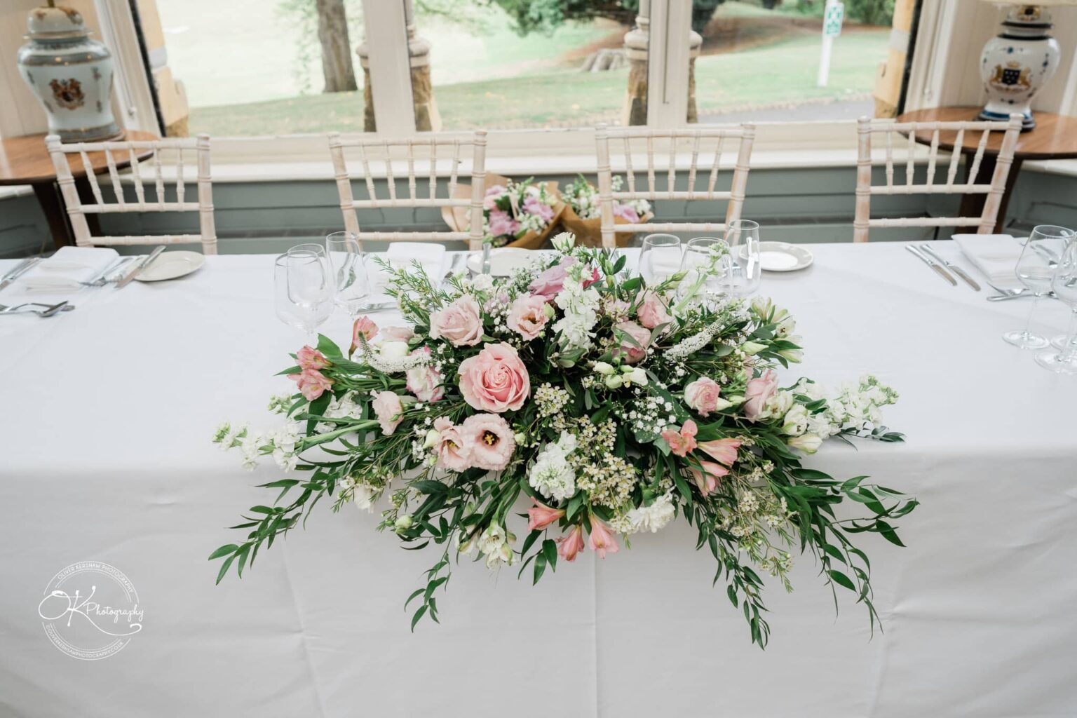 Ettington Park Hotel Wedding Photography Rectangular table with white tablecloth, decorated with a floral arrangement of pink and white flowers, surrounded by wooden chairs set for a meal, in a room with large windows overlooking greenery.