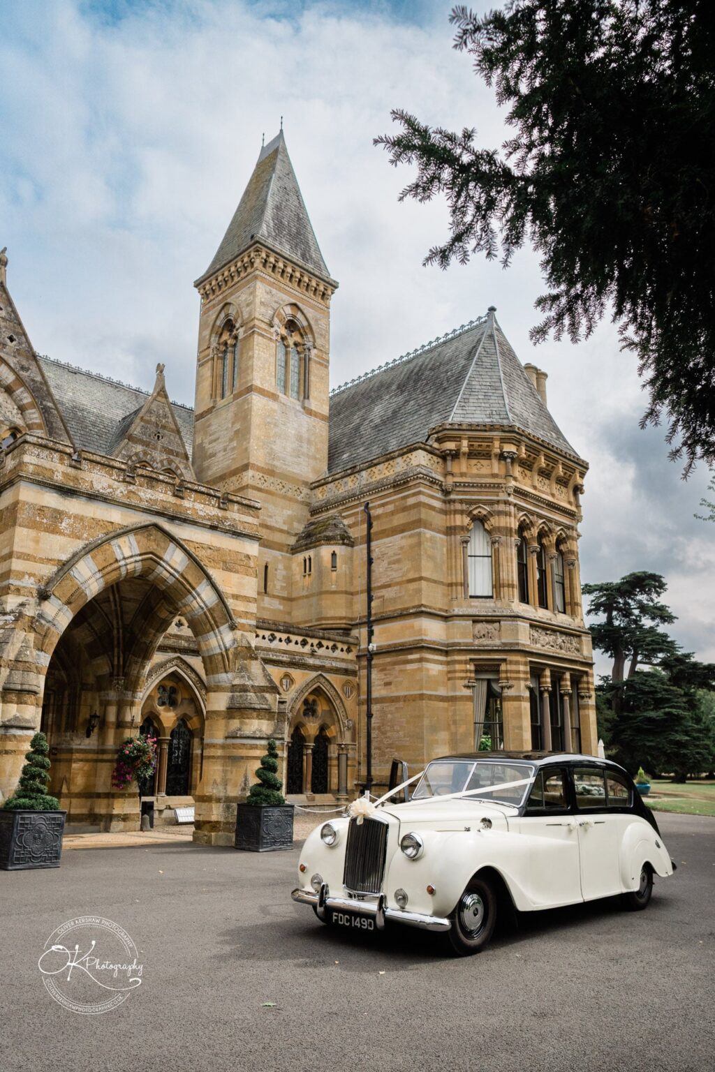 Ettington Park Hotel Wedding Photography A classic white car with a ribbon on the front bumper parked in front of a large, historic building with arched windows and intricate stonework.