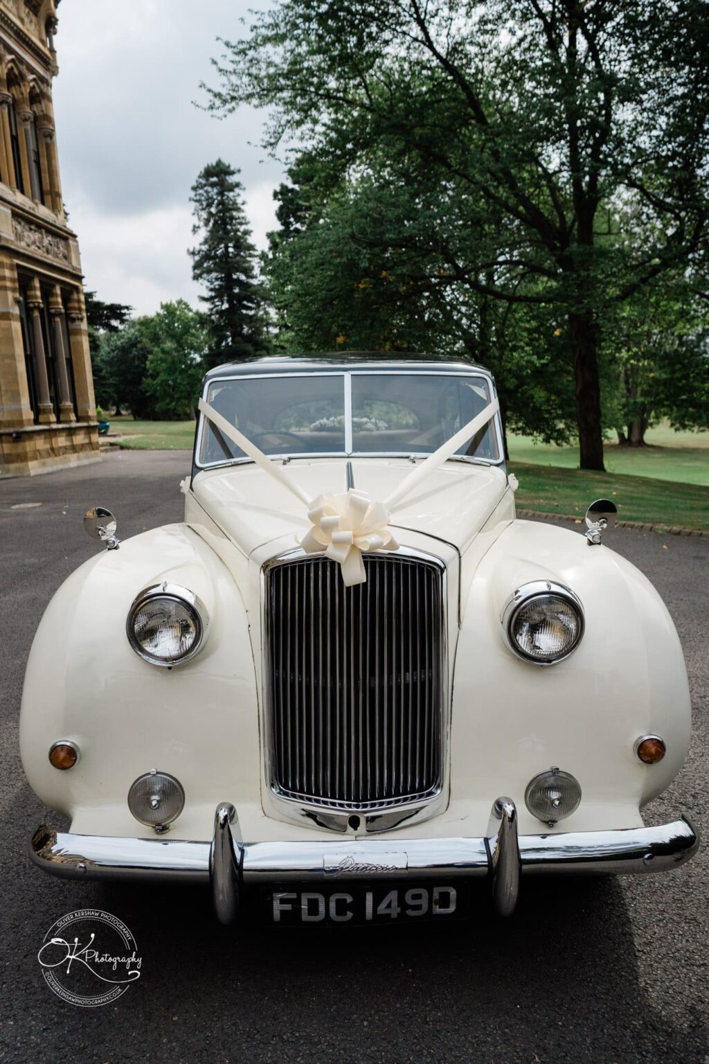 Ettington Park Hotel Wedding Photography Front view of a vintage white car decorated with a large ribbon bow on the bonnet, parked outside an ornate building with trees in the background.