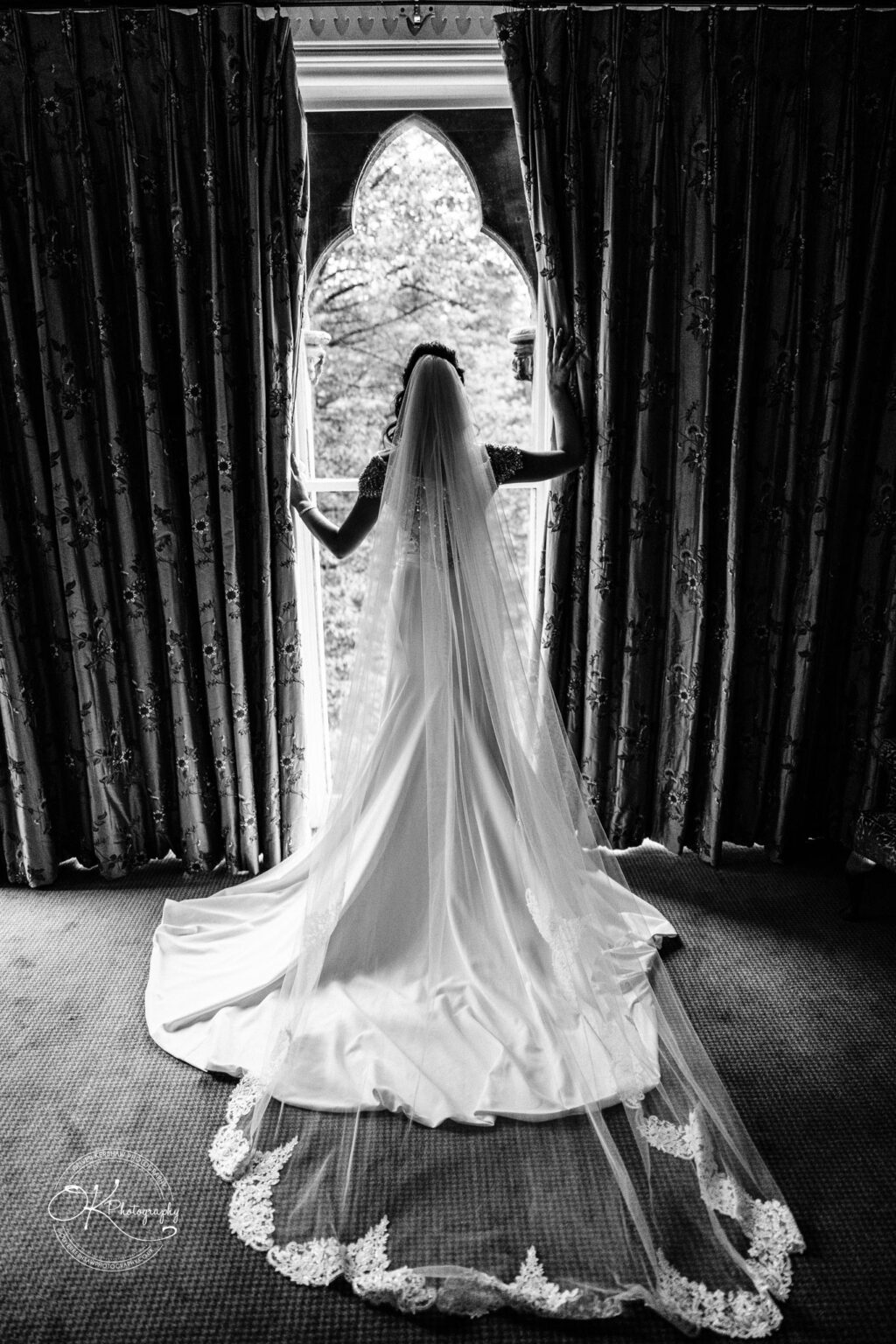 Ettington Park Hotel Wedding Photography Bride in a wedding gown and veil, standing in front of ornate curtains and looking out a window.