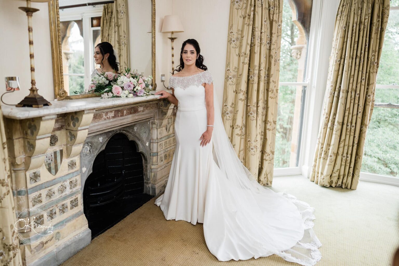 Ettington Park Hotel Wedding Photography Bride in an off-shoulder wedding dress with a long train, standing beside a decorated fireplace with a floral arrangement, looking at her reflection in the mirror.