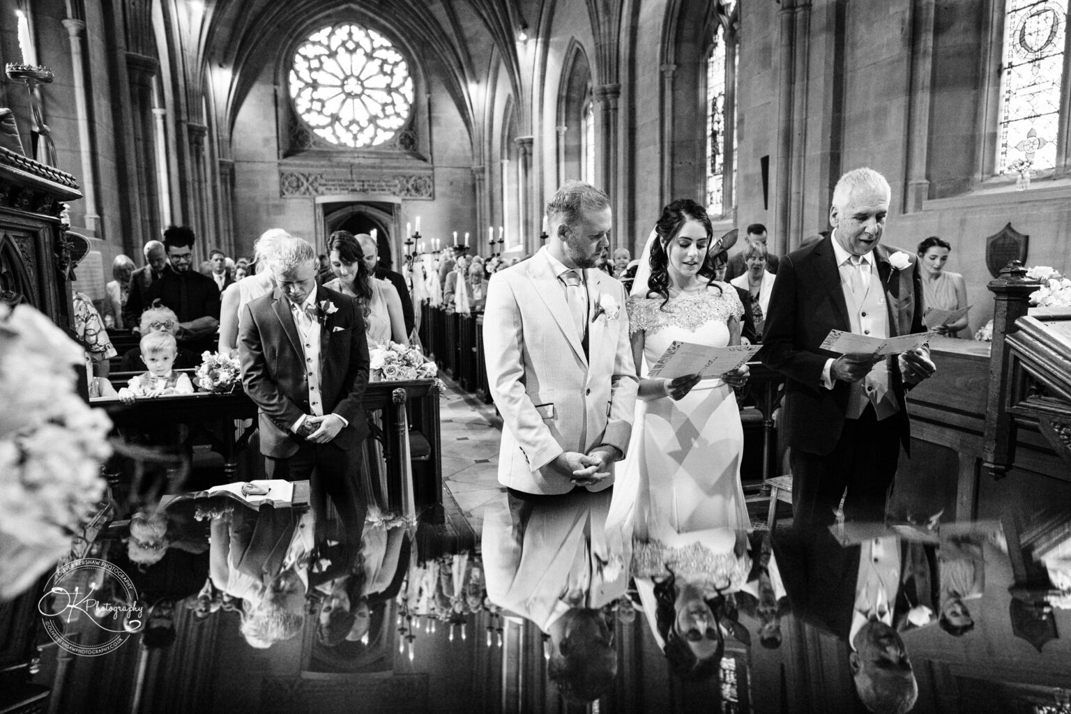 Ettington Park Hotel Wedding Photography A wedding ceremony inside a church with the bride and groom standing and holding papers alongside another man, a boy and guests seated in the background, surrounded by ornate architecture and stained glass windows.
