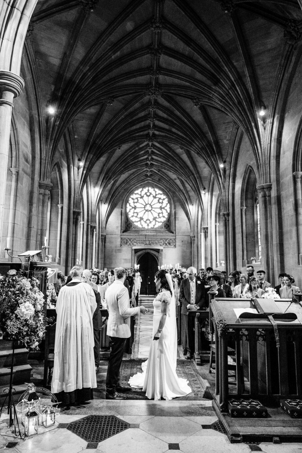 Ettington Park Hotel Wedding Photography Bride and groom standing at the altar in a gothic-style church, surrounded by guests and a priest conducting the ceremony.
