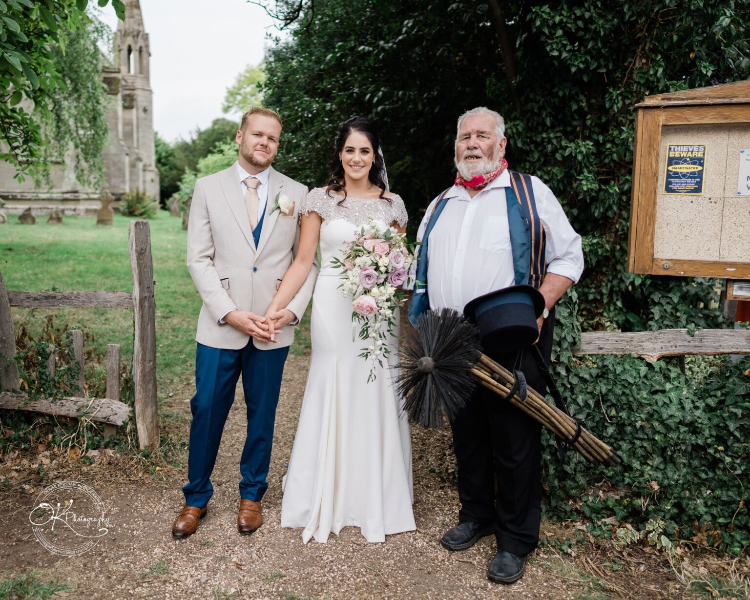 Ettington Park Hotel Wedding Photography Bride and groom standing with a chimney sweep holding his tools, near a church on a green grassy area.