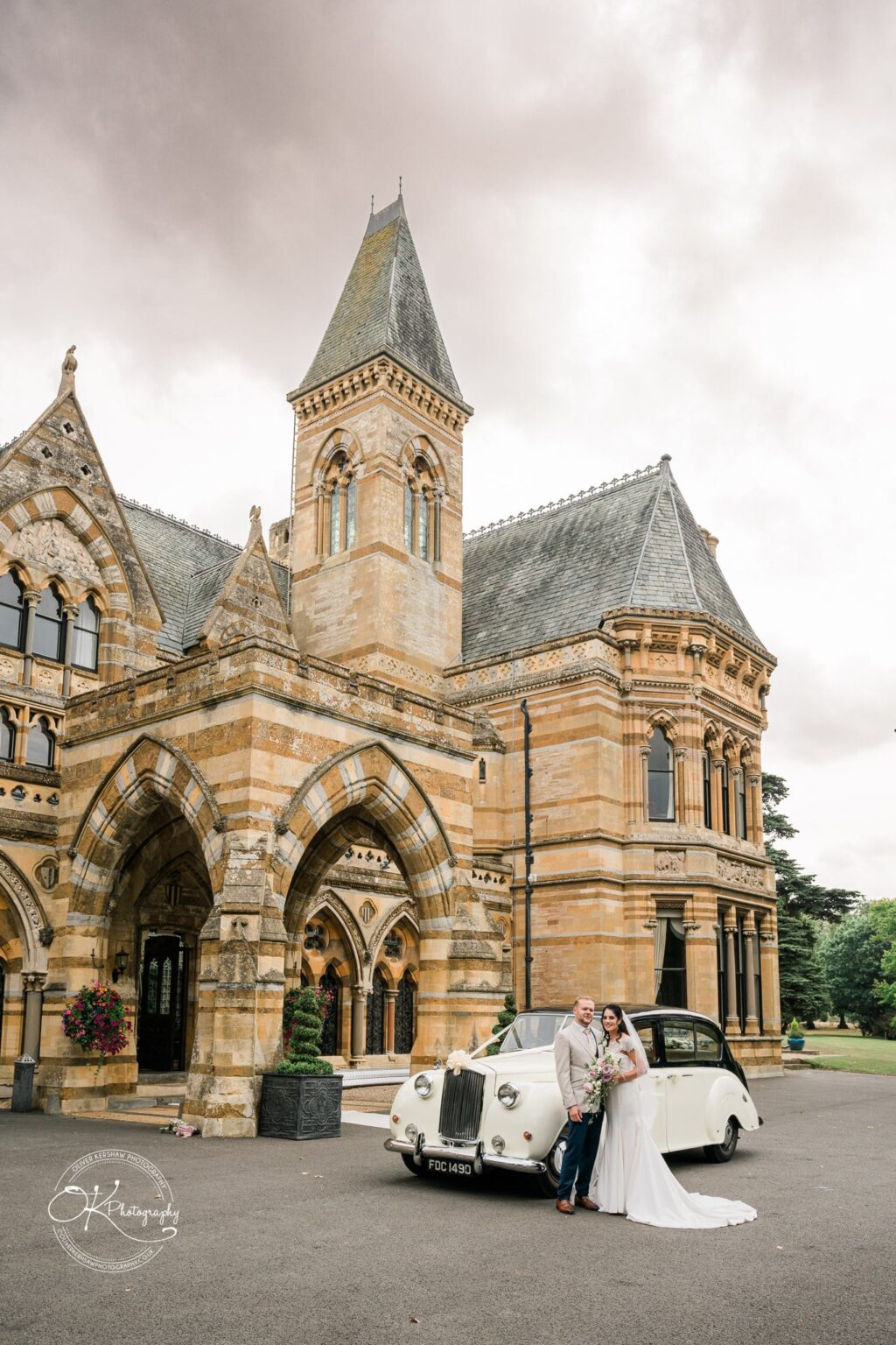 Ettington Park Hotel Wedding Photography Bride and groom standing in front of a vintage car outside a grand, historic building with Gothic architectural features.