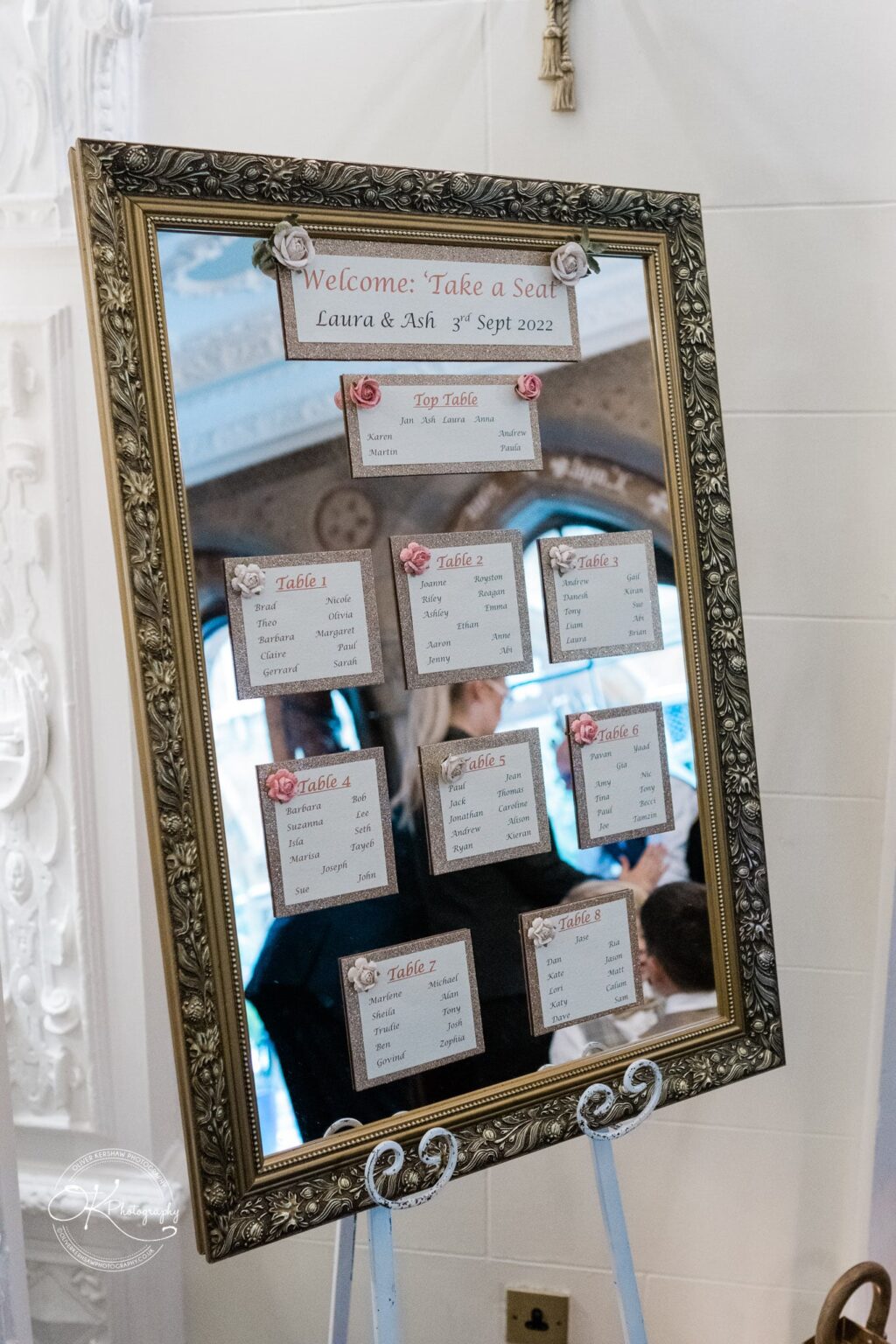 Ettington Park Hotel Wedding Photography A mirror wedding seating chart with a gold ornate frame, displaying the names of guests and their assigned tables. The sign at the top reads "Welcome: Take a Seat Laura & Ash, 3rd Sept 2022."
