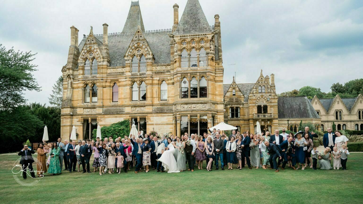 Ettington Park Hotel Wedding Photography A large group of people gathered on the lawn in front of a grand Victorian-style mansion for a wedding photo.