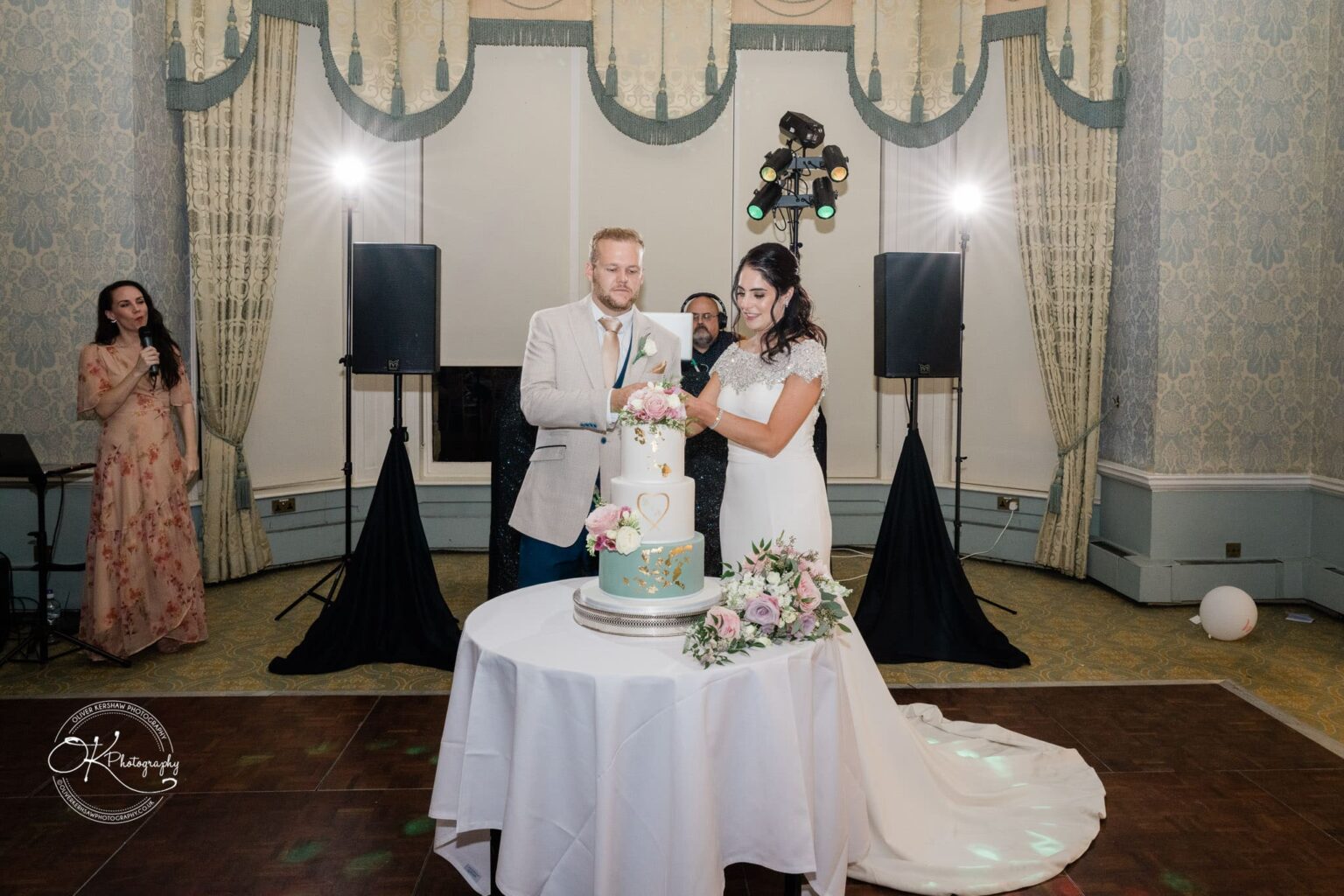 Ettington Park Hotel Wedding Photography Bride and groom cutting a three-tier wedding cake adorned with flowers and gold decorations, surrounded by a decorated banquet hall with onlookers in the background.